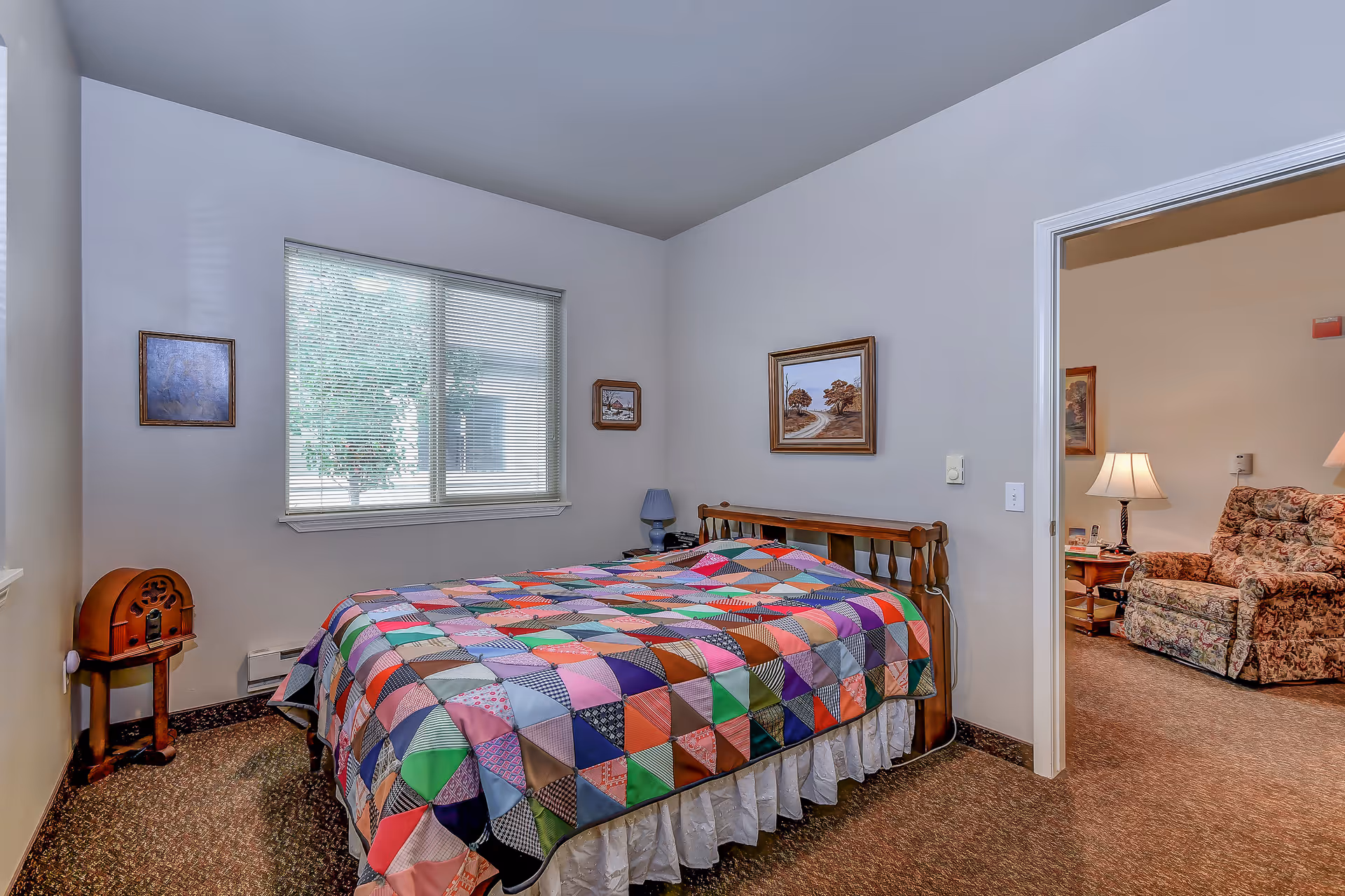 A bedroom with a wooden bed frame covered by a colorful patchwork quilt. The room has beige walls with three framed pictures hanging. A window with blinds lets in natural light. To the left of the bed is a small wooden side table with a vintage radio on it. Through an open doorway, a living room area with a floral armchair and a lamp on a side table is visible.