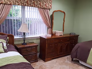 A bedroom with two twin beds, a wooden dresser with mirror and nightstand with a lamp beneath a window with vertical blinds and floral drapes.