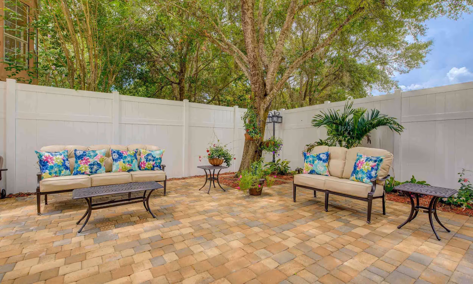 Outdoor patio area with two beige cushioned sofas adorned with colorful floral pillows, a black metal coffee table, and a small side table. The patio is paved with light brown bricks and surrounded by a white fence. There are several plants and a large tree providing shade in the corner.