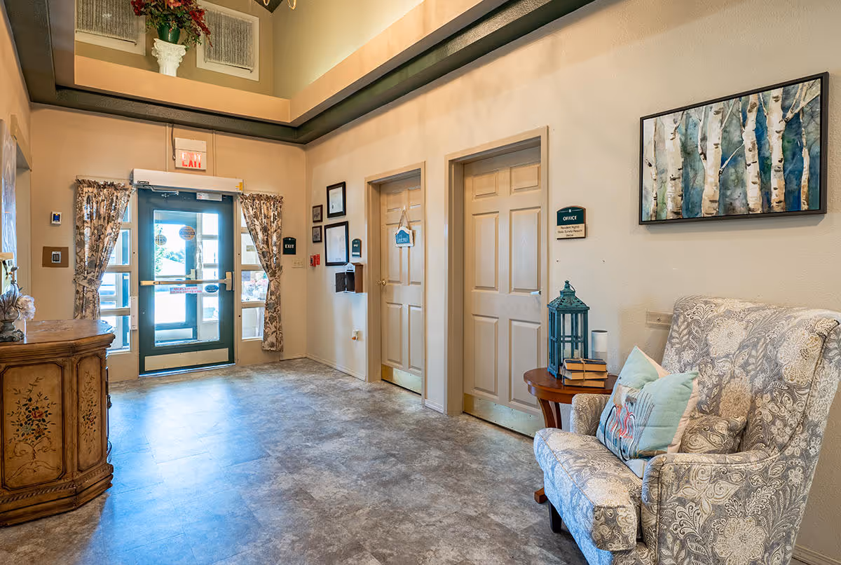 Interior view of a senior living facility hallway with a patterned armchair and side table with books and a lantern. There are two closed doors labeled 'Office' and 'Laundry', framed pictures on the wall, and a glass exit door with floral curtains letting in natural light.