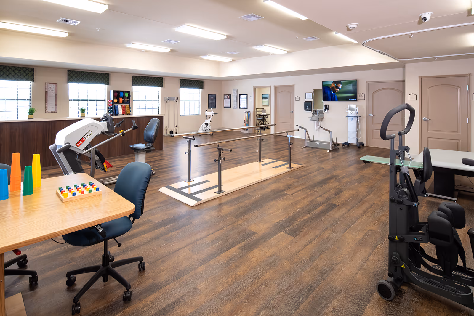 Spacious therapy and exercise room with parallel bars, various rehab machines, a table with chairs and activity supplies.