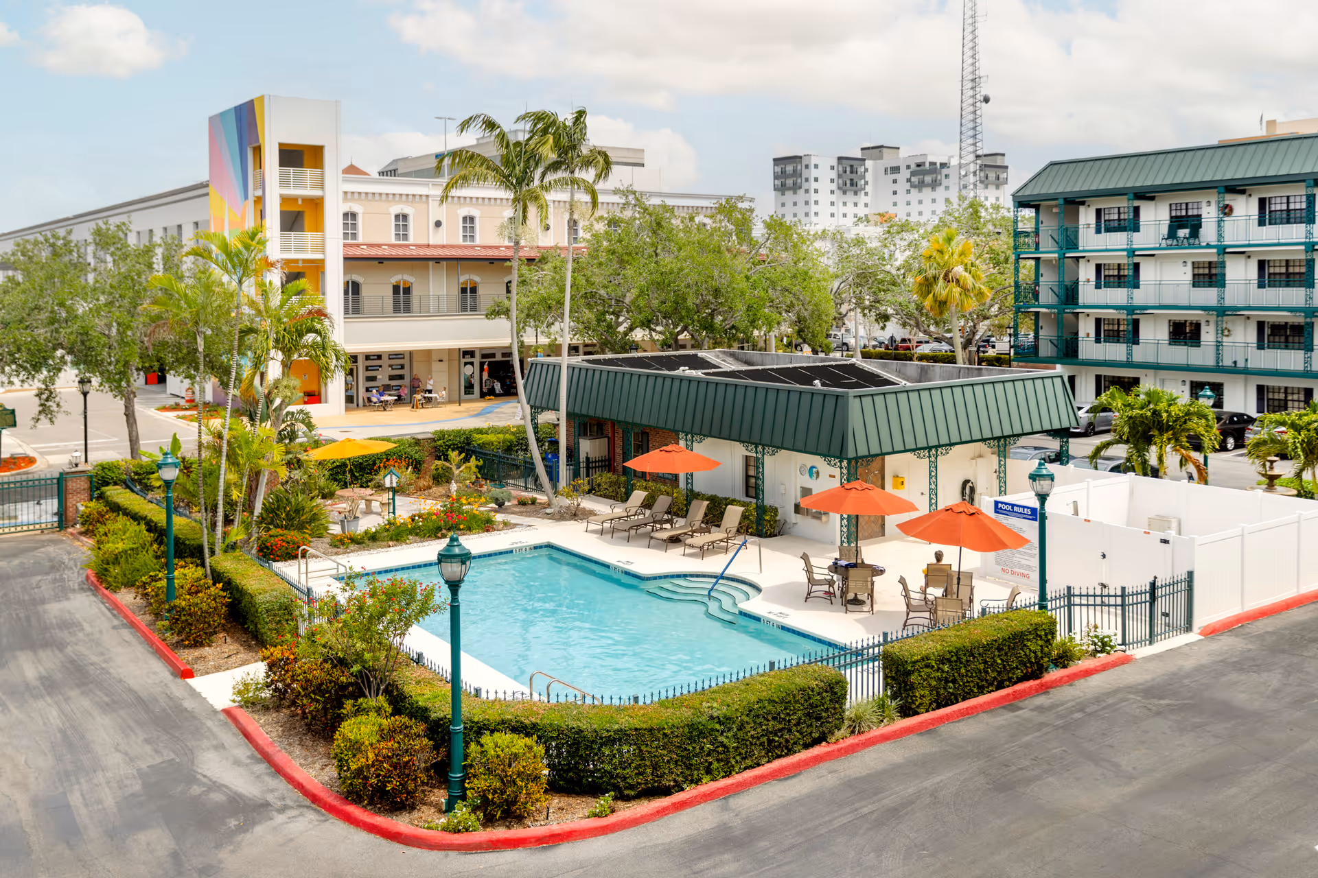 Outdoor swimming pool area with lounge chairs and tables shaded by orange umbrellas, surrounded by greenery and fencing. Behind the pool is a building with a green roof and a sign displaying pool rules. In the background, there are multi-story buildings, palm trees, and a partly cloudy sky.