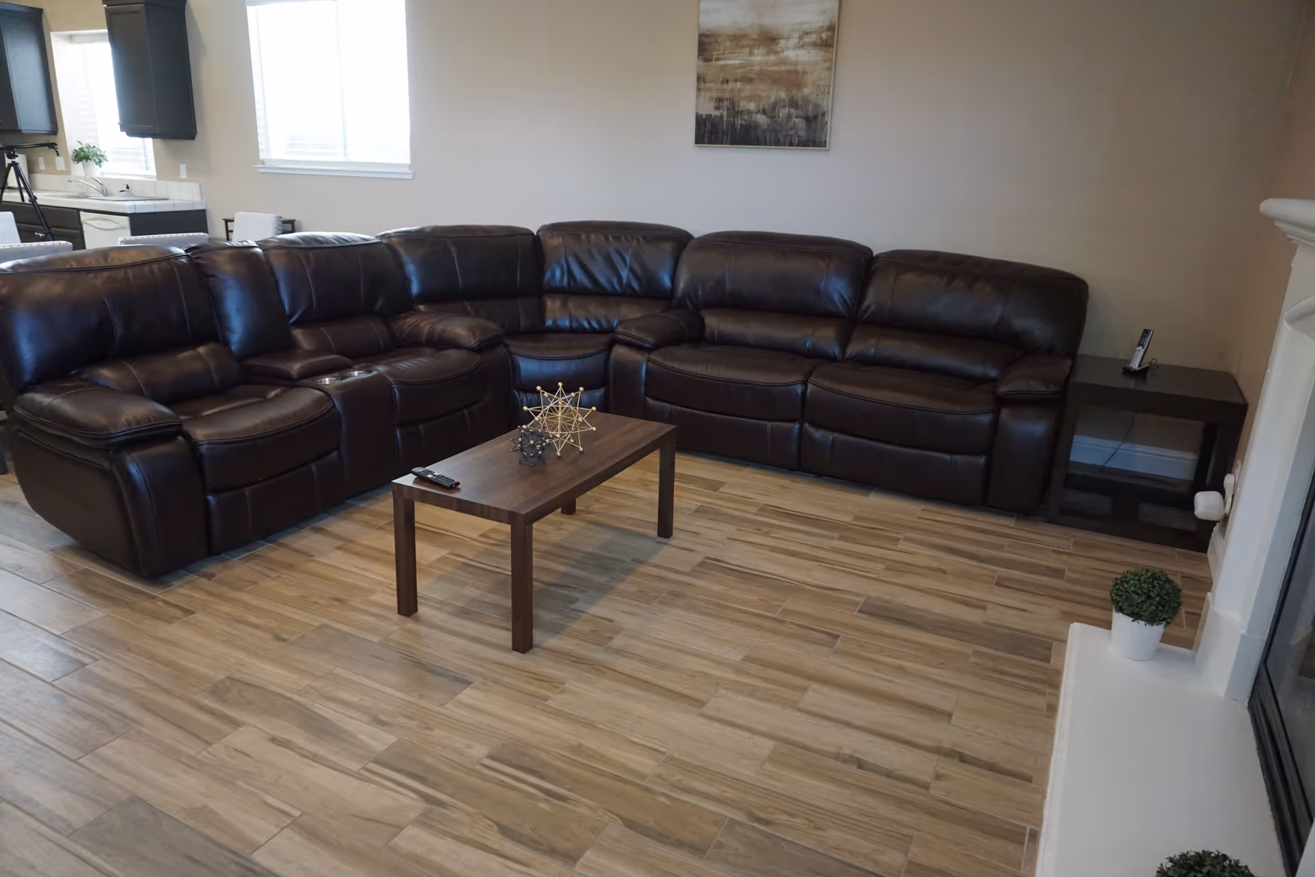A living room with a large dark brown leather sectional sofa arranged in an L-shape. In front of the sofa is a wooden coffee table with decorative geometric sculptures and a remote control on it. The room has light wood-look tile flooring, a small side table with a phone, a fireplace with a small potted plant on the hearth, and a window letting in natural light. A kitchen area is partially visible in the background.