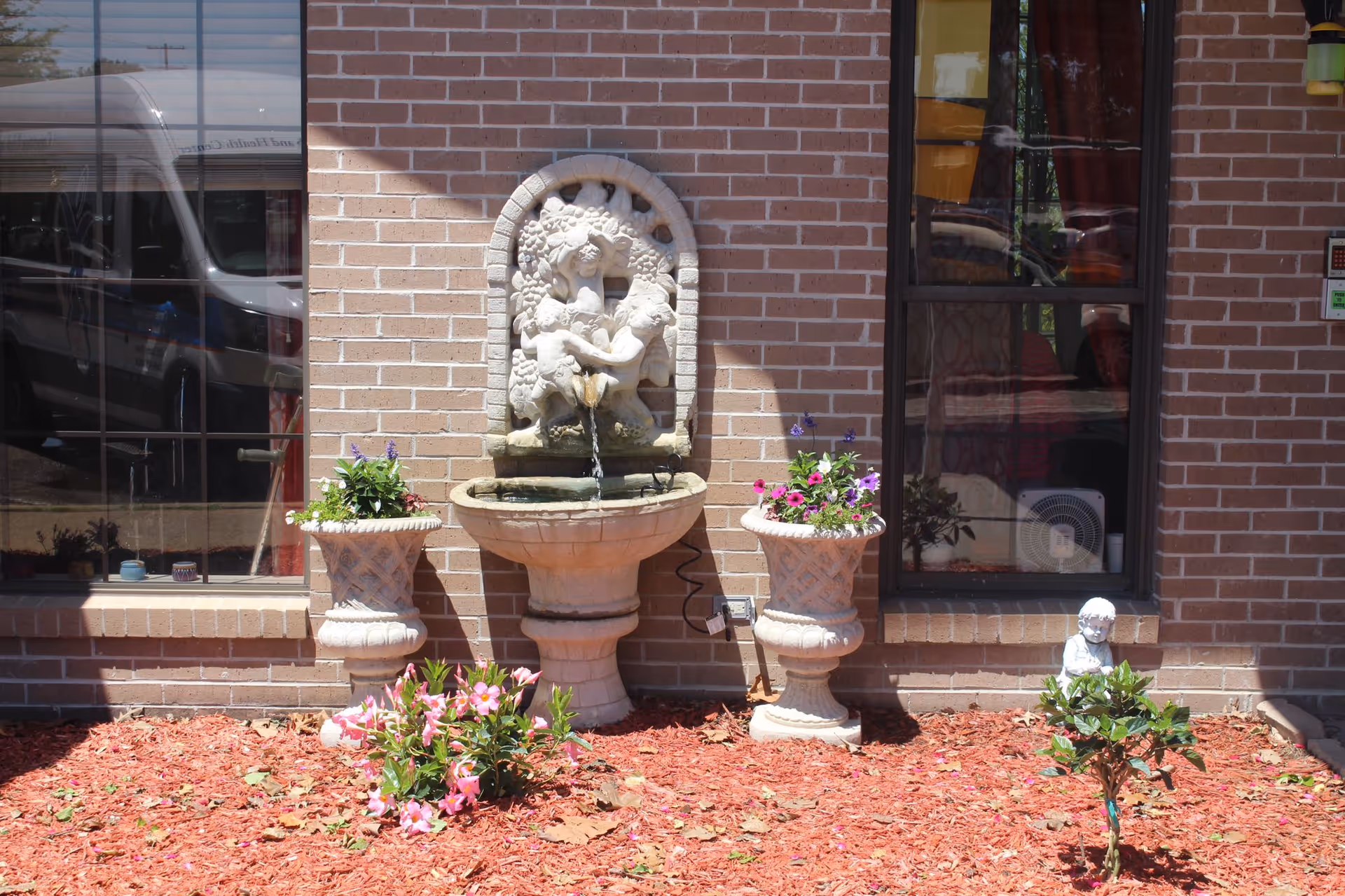 Outdoor garden area with a decorative stone fountain featuring cherubs mounted on a brick wall. Two large stone planters with flowers flank the fountain, and a small statue of a child is placed near a small shrub in the mulch bed. Windows of the building reflect the surroundings.