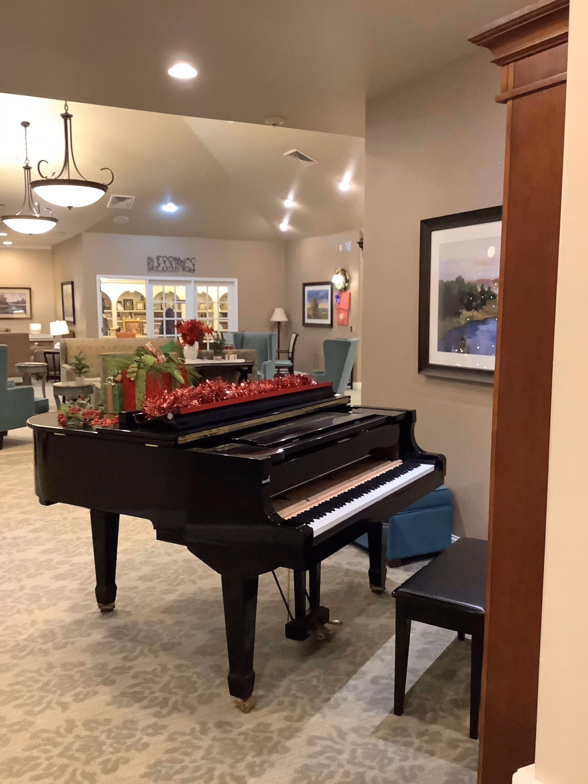 Interior view of a senior living facility lounge area featuring a black grand piano decorated with red garland and holiday decorations. The room has beige walls and carpet, with several chairs and tables arranged around the space. There are framed pictures on the walls and ceiling lights providing illumination.