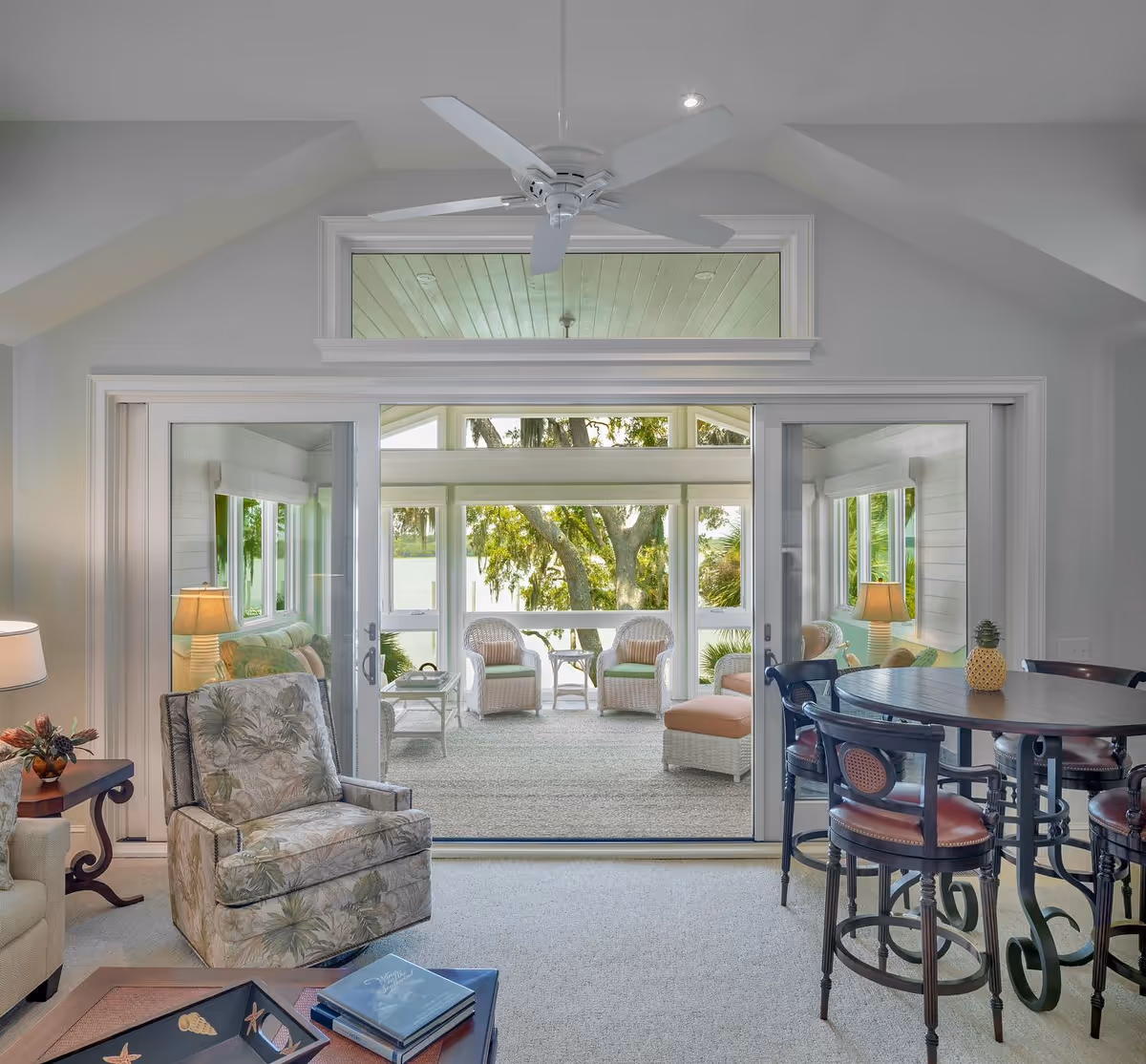 Bright living room with armchairs and a round dining table, sliding glass doors opening to a sunroom with wicker seating overlooking trees and water.