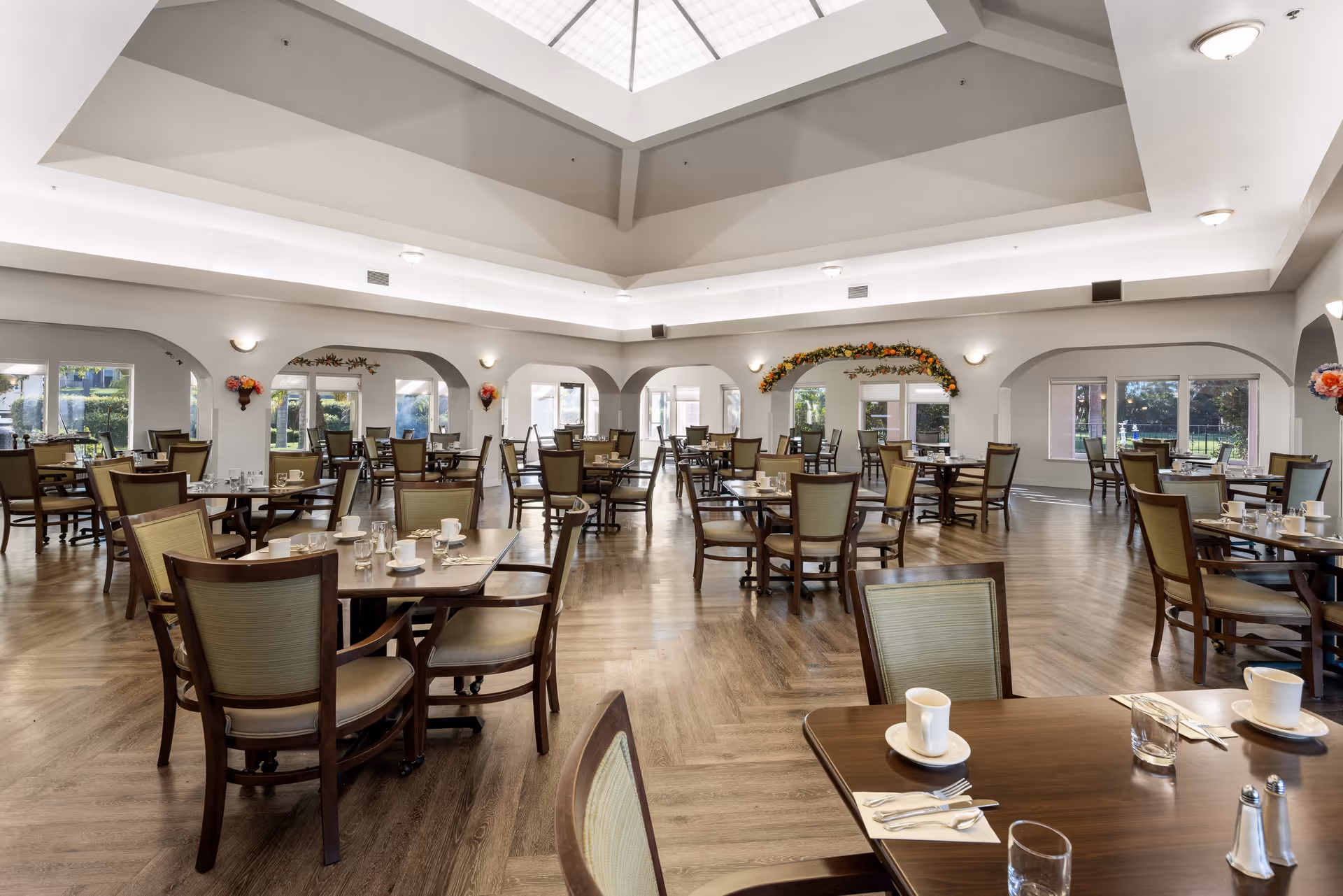 A spacious senior living community dining room with multiple wooden tables and chairs arranged neatly. Each table is set with white cups, saucers, glasses, and silverware. The room features large windows allowing natural light, a high ceiling with a skylight, and decorative floral arrangements on the walls.