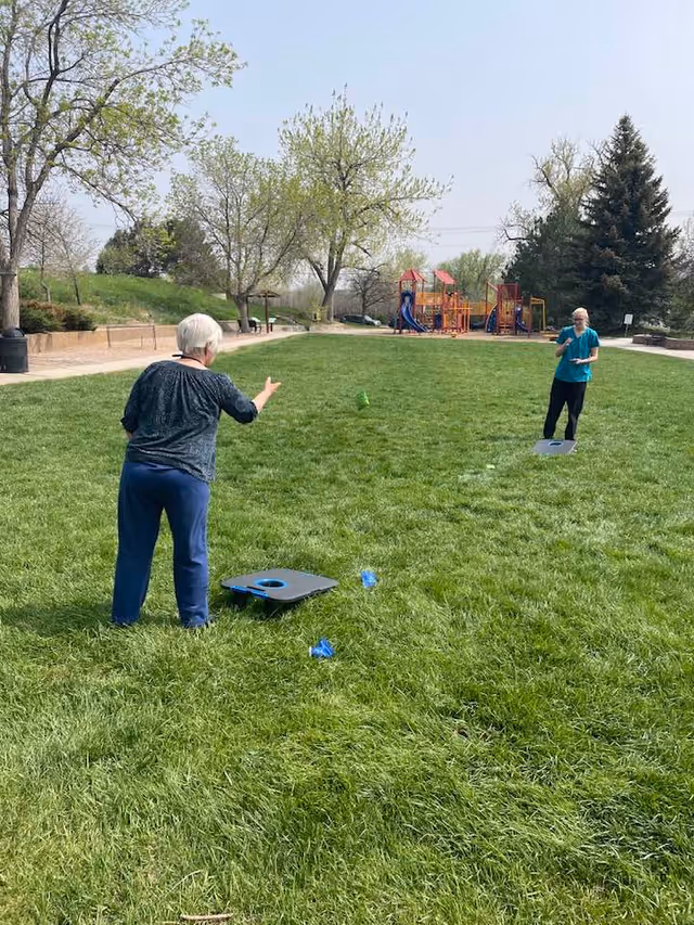 Two elderly women playing cornhole on a grassy field in a park with trees and a playground in the background on a clear day.