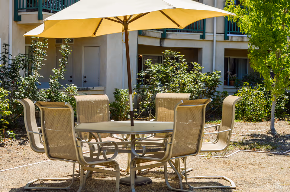 Outdoor patio area with a round table and six beige chairs under a large yellow umbrella, surrounded by greenery and part of a building with balconies in the background.