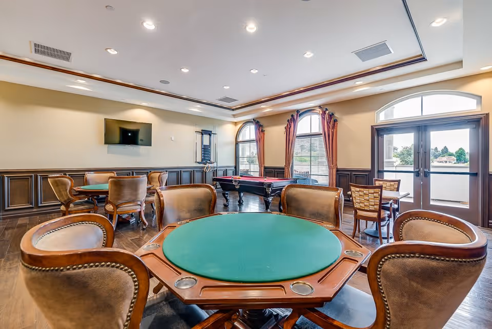 A spacious senior living common room with multiple card tables surrounded by leather chairs, a pool table in the background, large arched windows with curtains, and a wall-mounted TV. The room has wooden flooring and a door leading to an outdoor area with a view of trees and buildings.