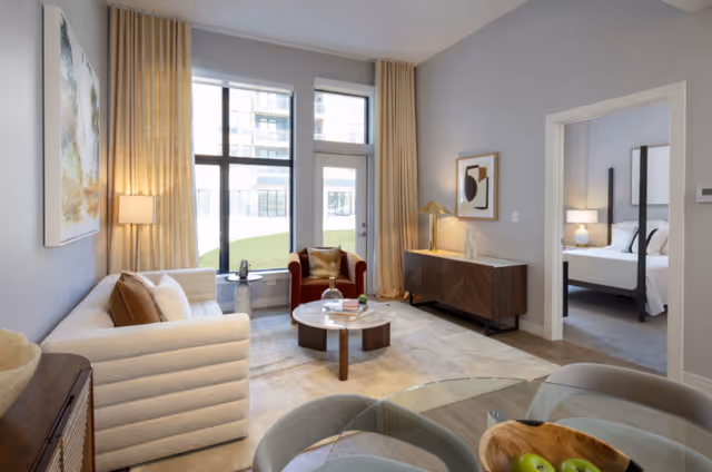 A modern living room with a white textured sofa, a red armchair, a round wooden coffee table, and a wooden sideboard with a lamp and decorative items. Large windows with beige curtains let in natural light, and a doorway leads to a bedroom with a bed and nightstand visible. A glass dining table with chairs and a bowl of green apples is in the foreground.