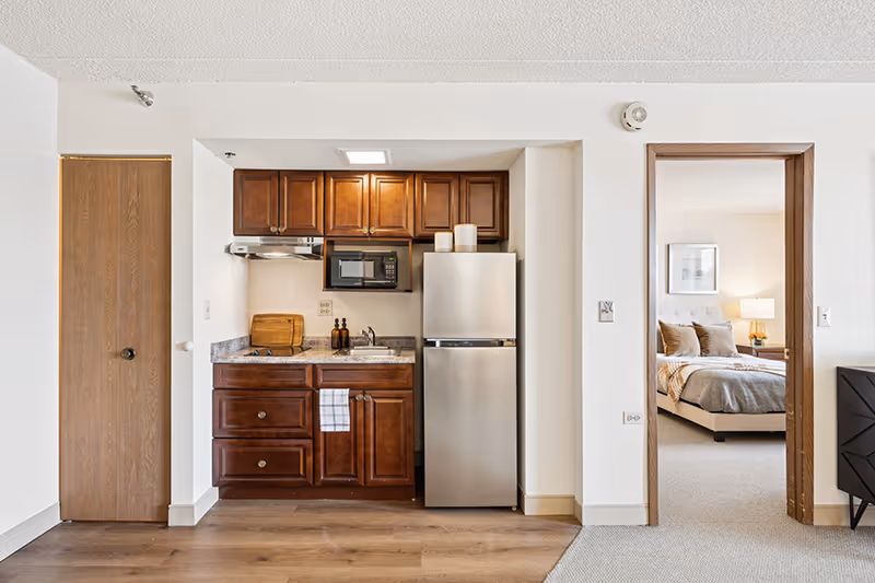 A compact kitchen area with wooden cabinets, a stainless steel refrigerator, a microwave, and a small sink. To the right, an open doorway reveals a bedroom with a bed, pillows, a bedside table, and a lamp. The floor is a combination of wood and carpet.