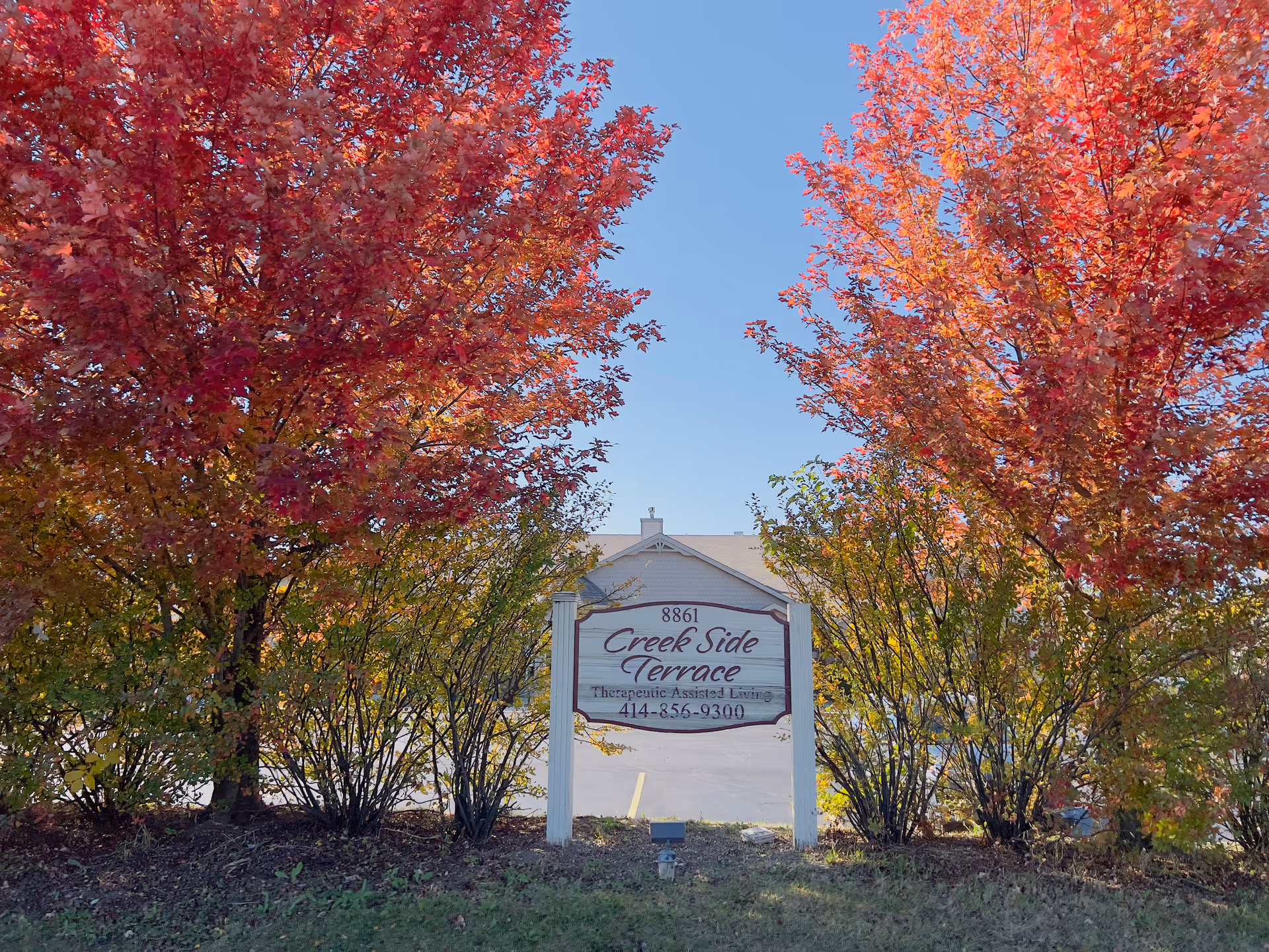 A white wooden sign for Creek Side Terrace Therapeutic Assisted Living with the phone number 414-856-9300, surrounded by vibrant red and orange autumn trees and bushes under a clear blue sky.