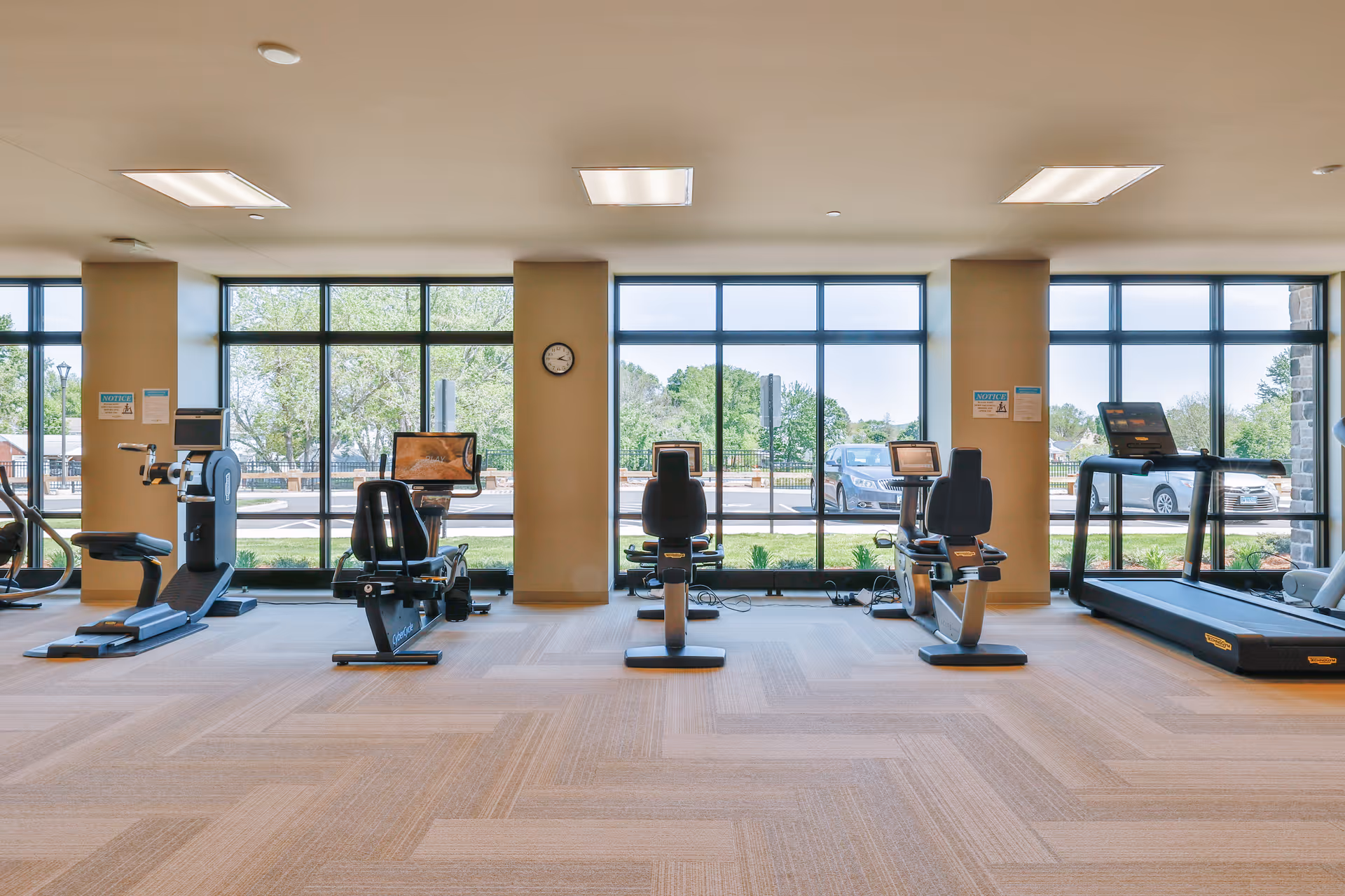 A bright fitness room with large windows showing an outdoor view. The room contains various exercise equipment including stationary bikes, a treadmill, and other cardio machines. The floor is carpeted and the walls are light-colored with a clock mounted on one pillar.