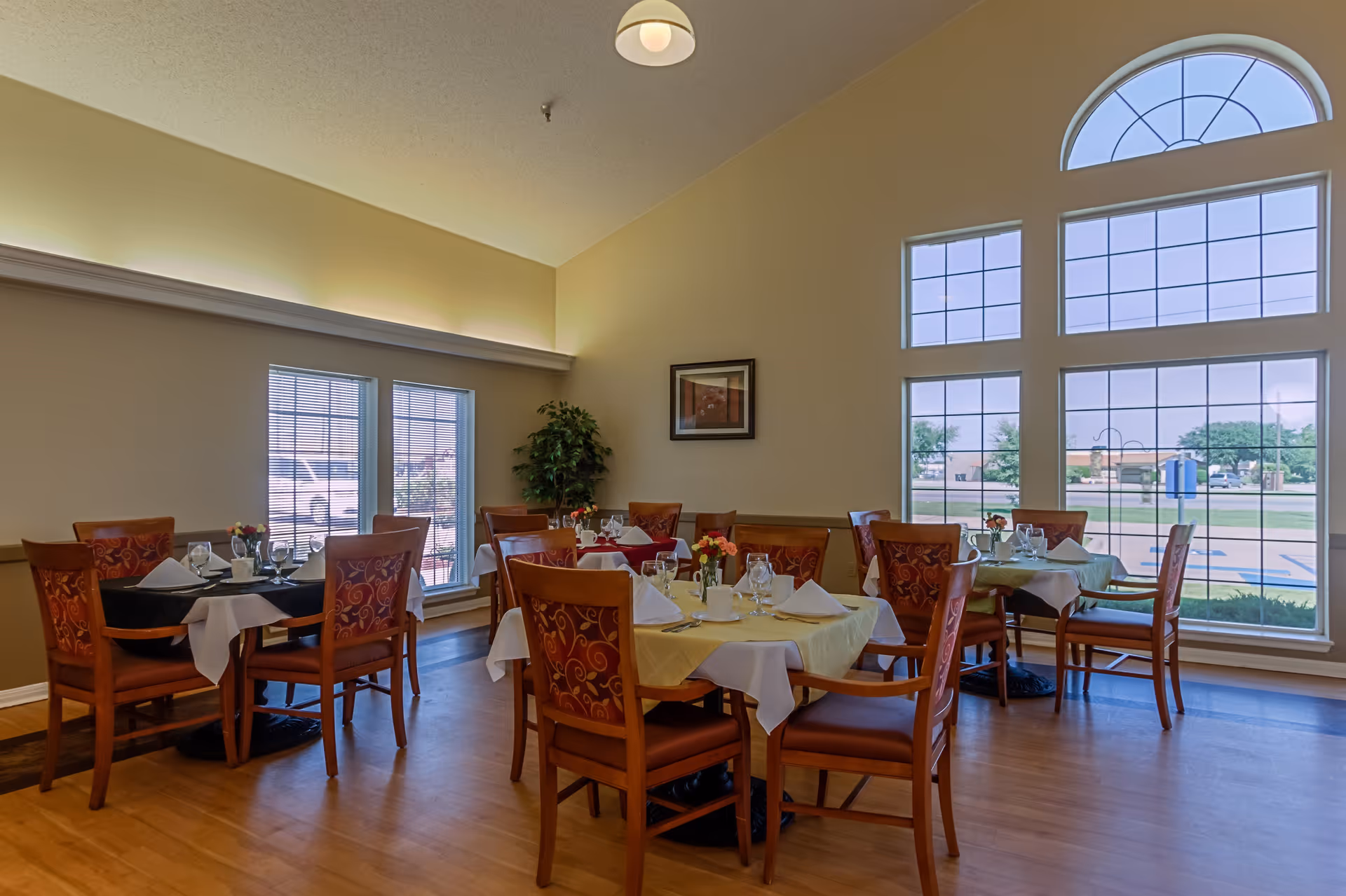 A dining room in a senior living facility with several wooden tables and chairs. The tables are set with white napkins, glassware, and small flower arrangements. Large windows with decorative panes allow natural light to fill the room, and there is a framed picture on the beige wall.