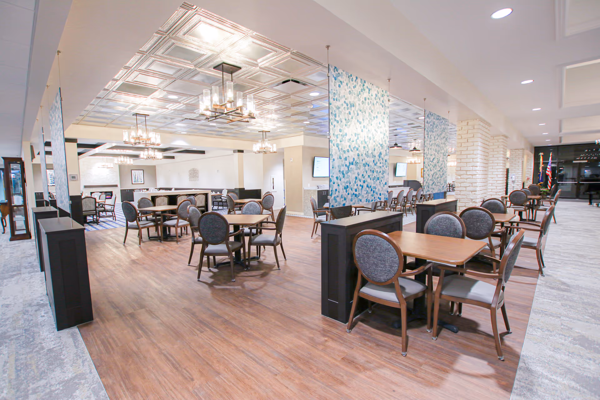 A spacious and well-lit dining area with multiple wooden tables and cushioned chairs arranged neatly. The room features a decorative ceiling with modern chandeliers, light wood flooring, and partitions with blue patterned panels. There are large windows and a glass door visible in the background, along with American flags near the entrance.