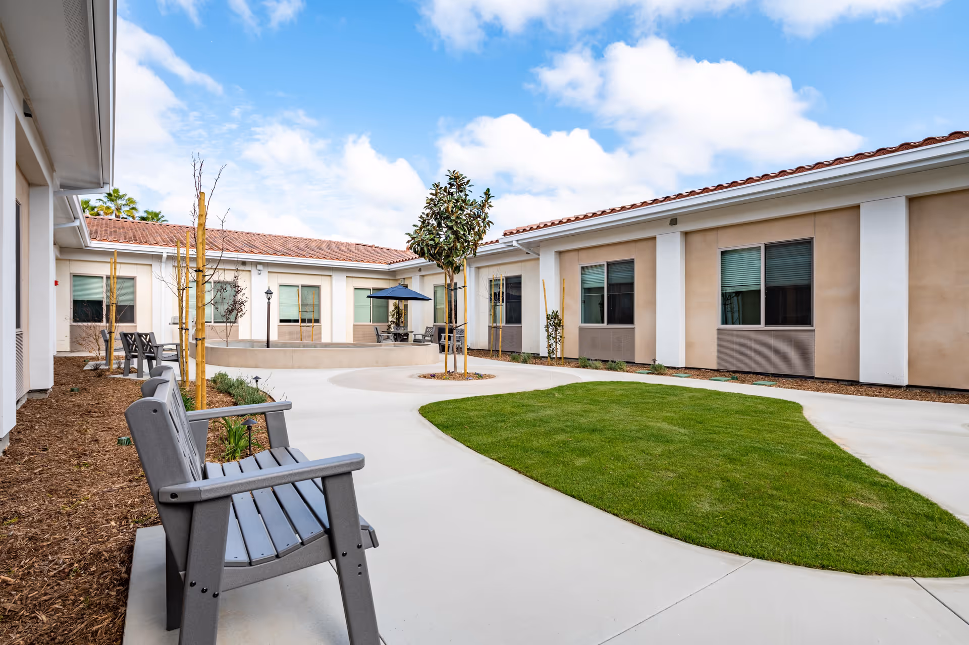Outdoor courtyard area at Park View Estates Assisted Living & Memory Care featuring a paved walkway, green lawn, young trees, and several gray benches. The building surrounding the courtyard has beige and white walls with windows and a red-tiled roof under a partly cloudy blue sky.