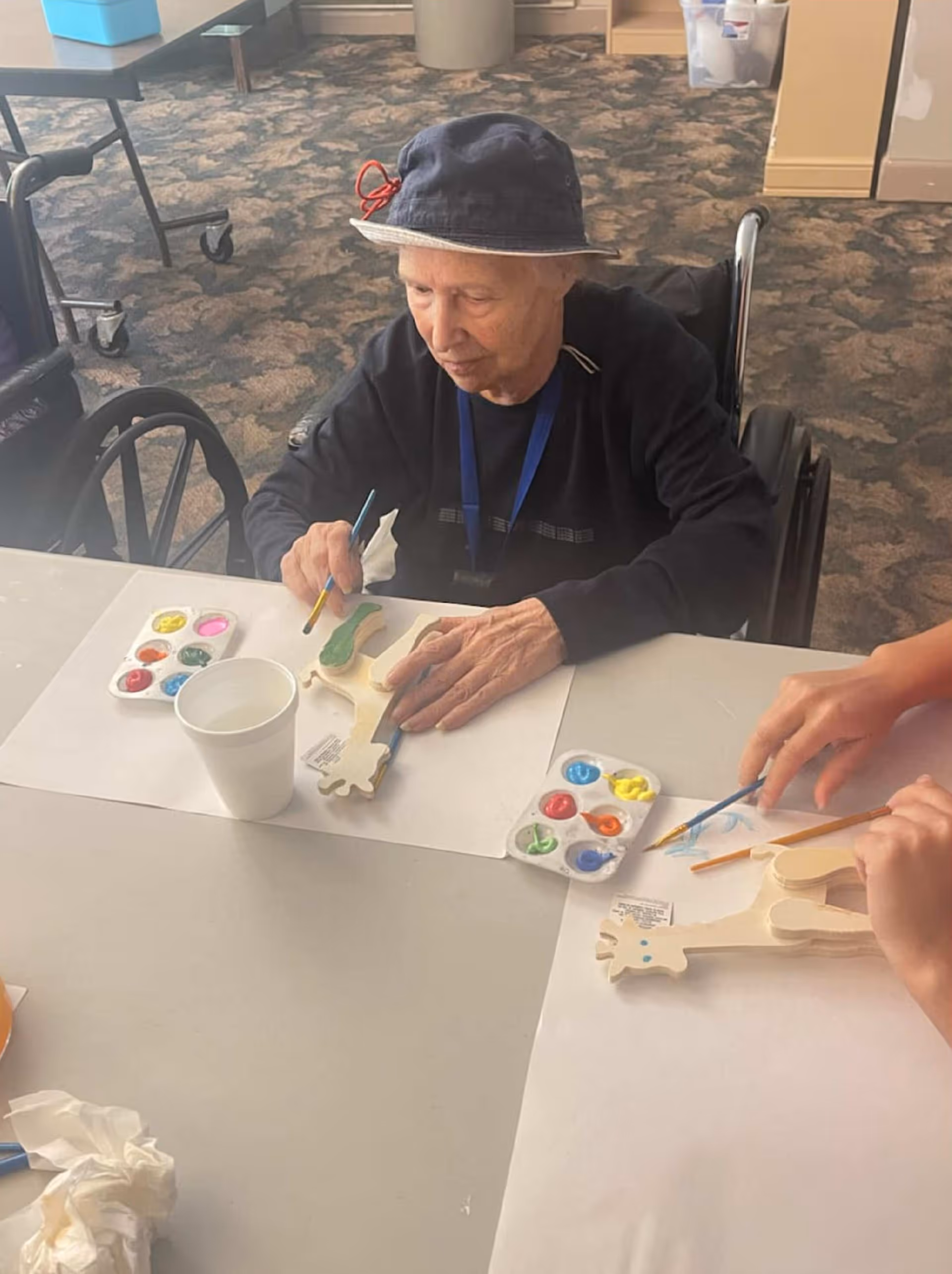 An elderly person in a wheelchair wearing a dark hat and sweater is painting a wooden giraffe figure at a table. There are paint palettes with various colors and a white cup on the table. Another person is also painting a similar wooden giraffe figure nearby.