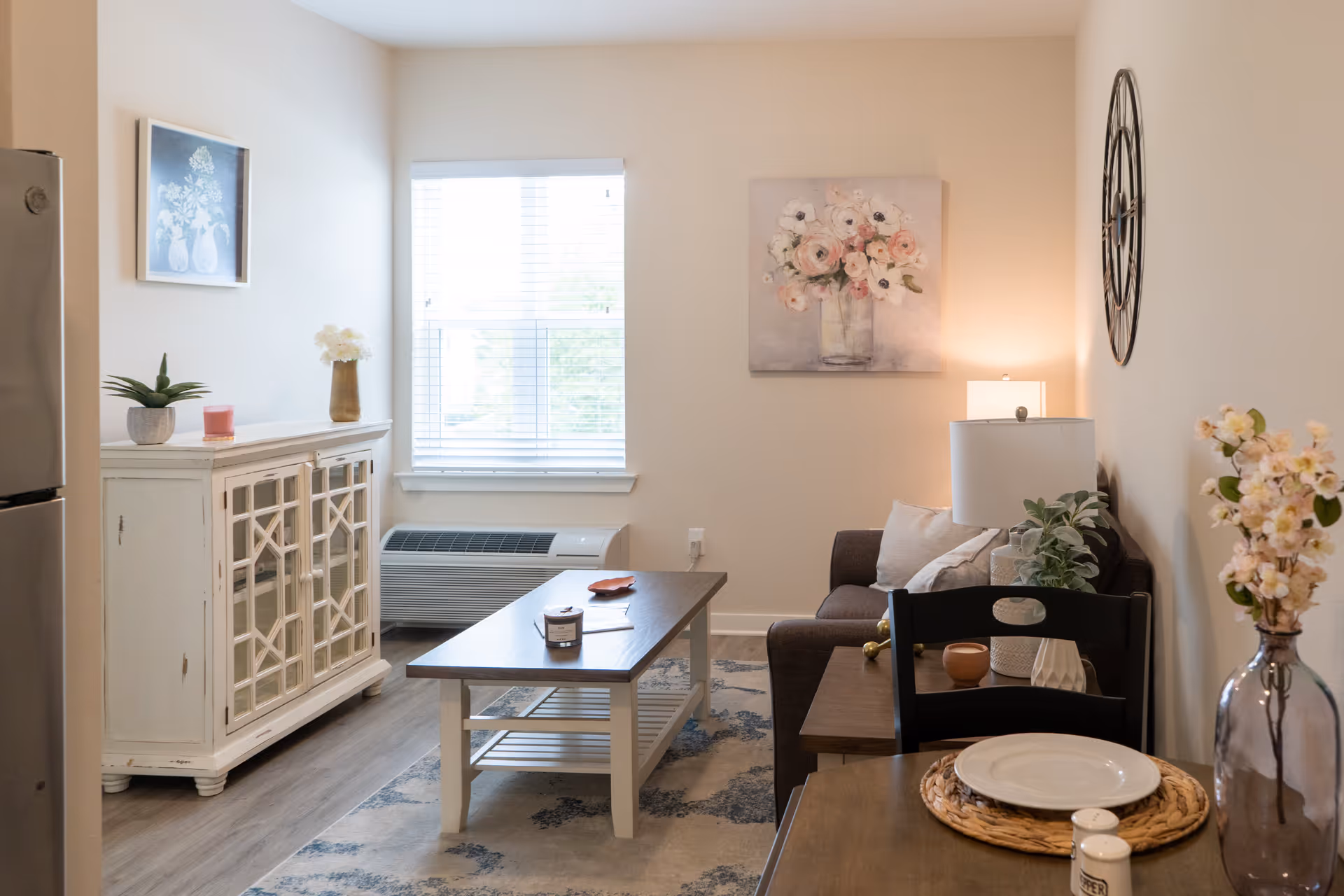 A cozy living room area with a brown sofa, a wooden coffee table, and a white cabinet. The room has light-colored walls with floral artwork and a large decorative clock. There is a window with white blinds letting in natural light, and a small dining table set with a plate and salt and pepper shakers. Various plants and decorative items are placed around the room.