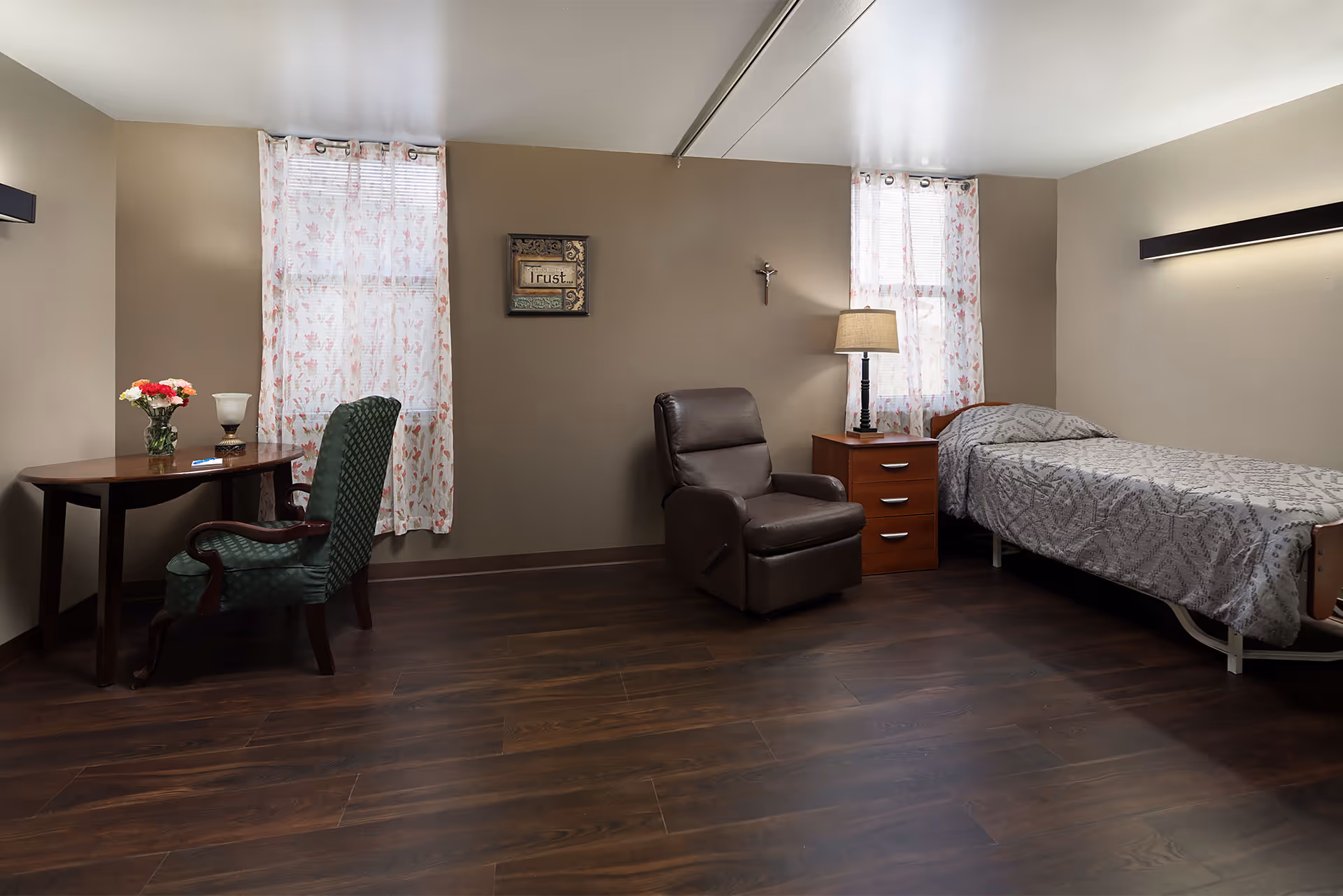 A simple, clean bedroom in a skilled nursing facility featuring a single bed with a patterned gray bedspread, a wooden nightstand with a lamp, a brown recliner chair, a wooden table with a green upholstered chair, and two windows with floral curtains letting in natural light. The walls are beige and the floor is dark wood.