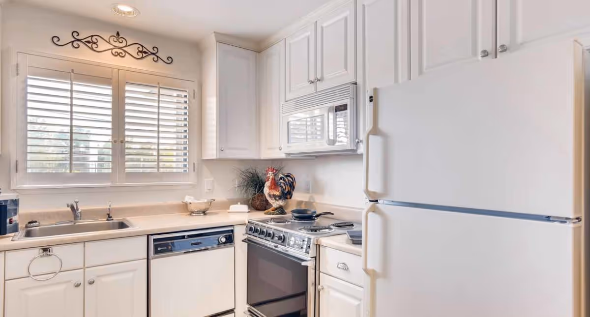 A bright kitchen with white cabinets, a white refrigerator, a microwave above a stove with a frying pan, a dishwasher, a double sink under a window with white shutters, and decorative items including a rooster figurine and a plant on the countertop.