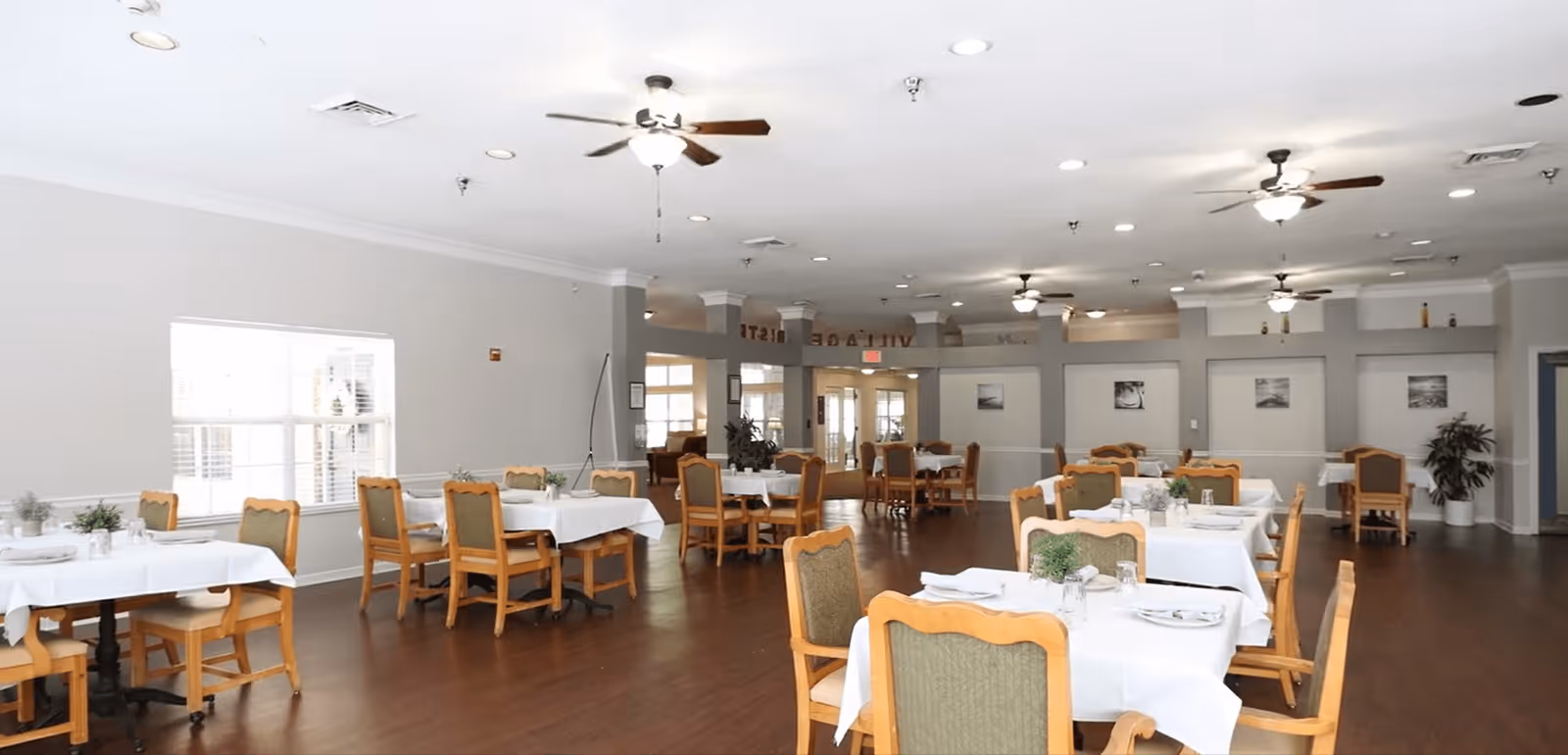A spacious dining room with multiple tables covered in white tablecloths, each set with plates, glasses, and napkins. Wooden chairs with green upholstery surround the tables. The room has large windows letting in natural light, ceiling fans, and framed pictures on the walls.