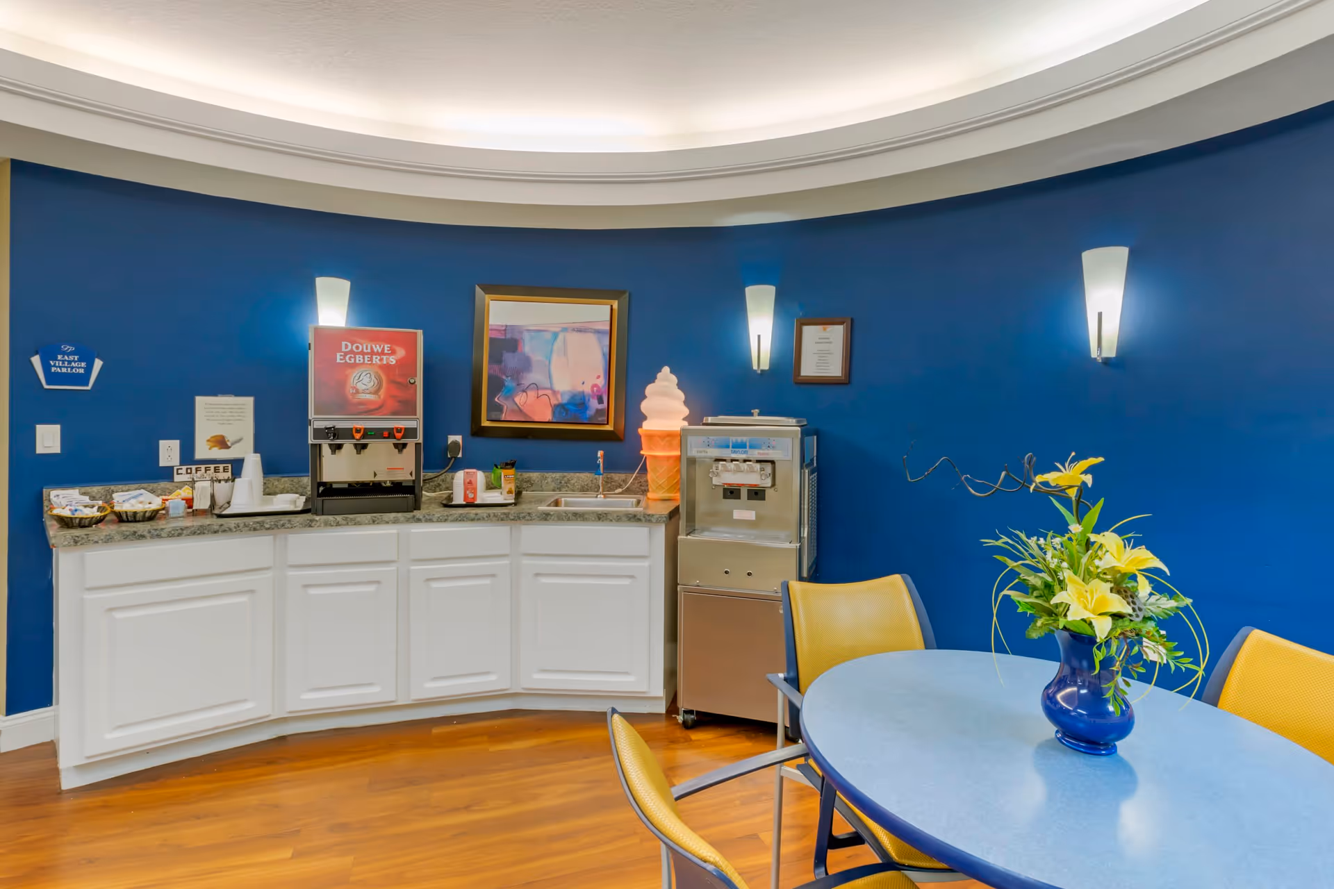 A coffee and ice cream station area with a blue wall background. The station includes a Douwe Egberts coffee dispenser, an ice cream machine with a large ice cream cone decoration on top, and a countertop with various coffee supplies. In front, there is a round table with yellow chairs and a blue vase with yellow flowers on it. The floor is wooden, and the ceiling has recessed lighting.