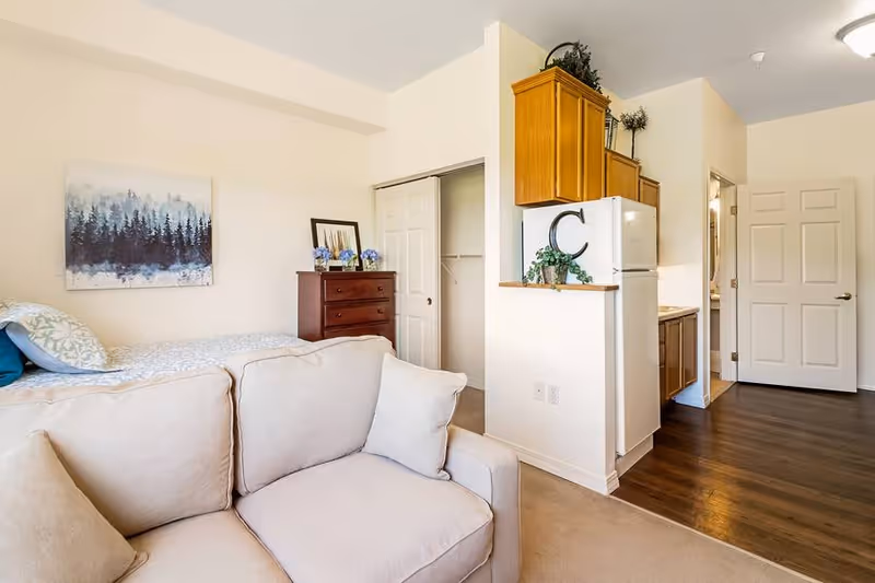 Interior view of a senior living facility room at Rexford Park showing a beige couch in the foreground, a bed with pillows and a forest-themed painting on the wall, a wooden dresser with decorative items, an open closet with sliding doors, and a small kitchen area with a refrigerator and wooden cabinets. The floor transitions from carpet to wood near the kitchen and hallway.