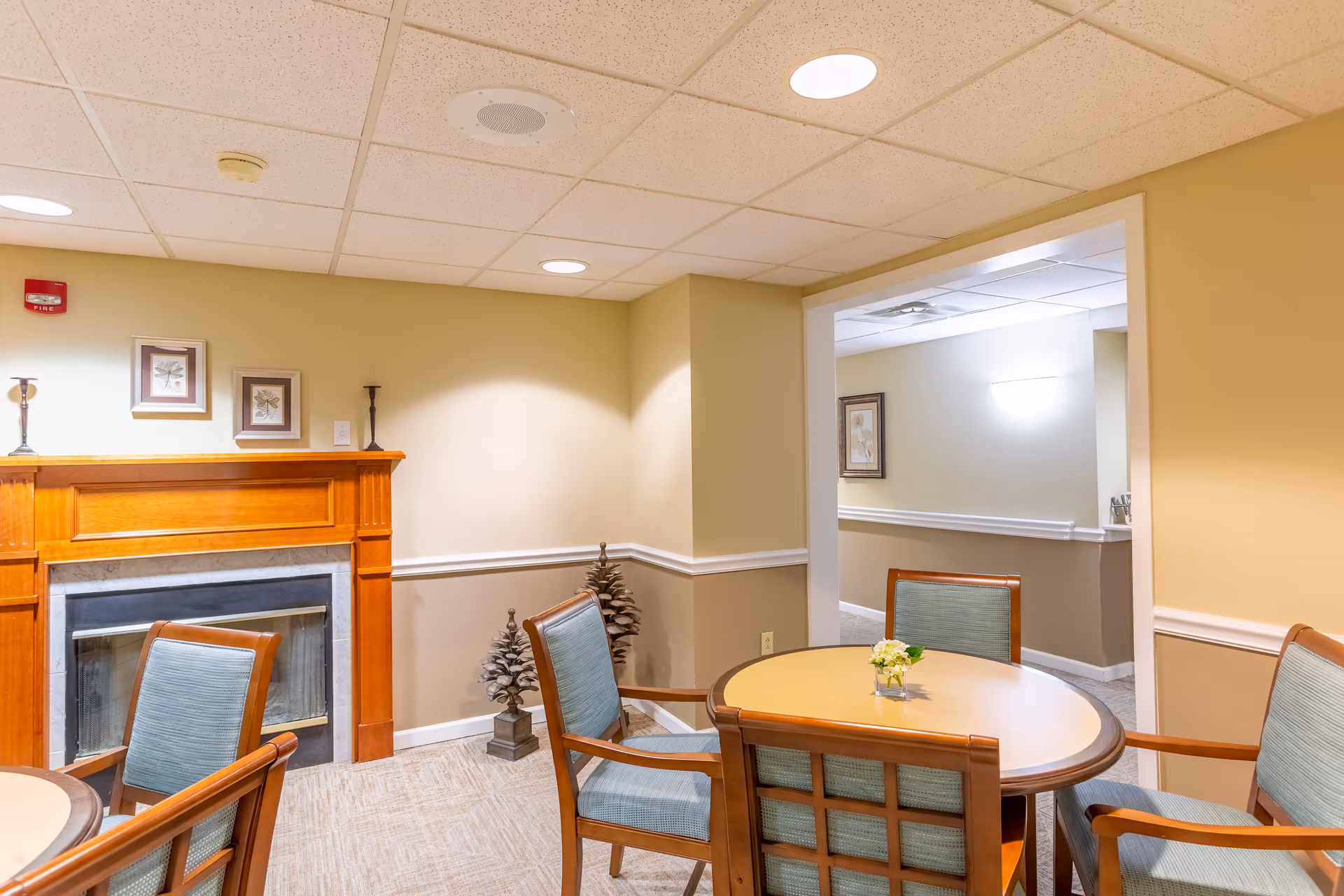 A cozy interior room with a round wooden table surrounded by four cushioned chairs. The walls are painted beige with white trim, and there is a wooden fireplace mantel with two framed pictures and candle holders on top. The ceiling has recessed lighting, and there is an open doorway leading to another room with similar decor.