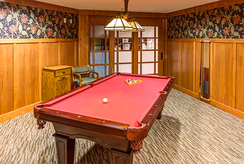 A room with a red felt pool table in the center, a wooden chair and small cabinet against the wall, floral wallpaper above wooden paneling, a carpeted floor, and a hanging light fixture above the pool table. Sliding glass doors are visible in the background.