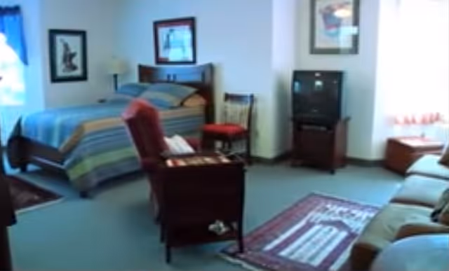 A cozy room with a bed covered in a striped blue and green blanket, a red armchair, a wooden chair with a red cushion, a TV on a stand, a sofa, and two framed pictures on the walls. The room has carpeted flooring and windows with curtains allowing natural light.