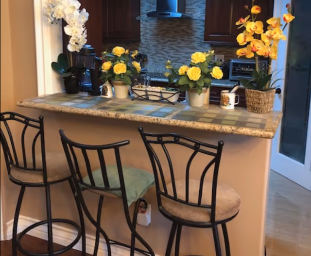 A kitchen counter with a tiled surface and three metal bar stools with cushioned seats. On the counter are several potted plants with yellow flowers and a white orchid, along with a coffee mug and a decorative tray. In the background, there are dark wooden kitchen cabinets, a stainless steel range hood, and a tiled backsplash.