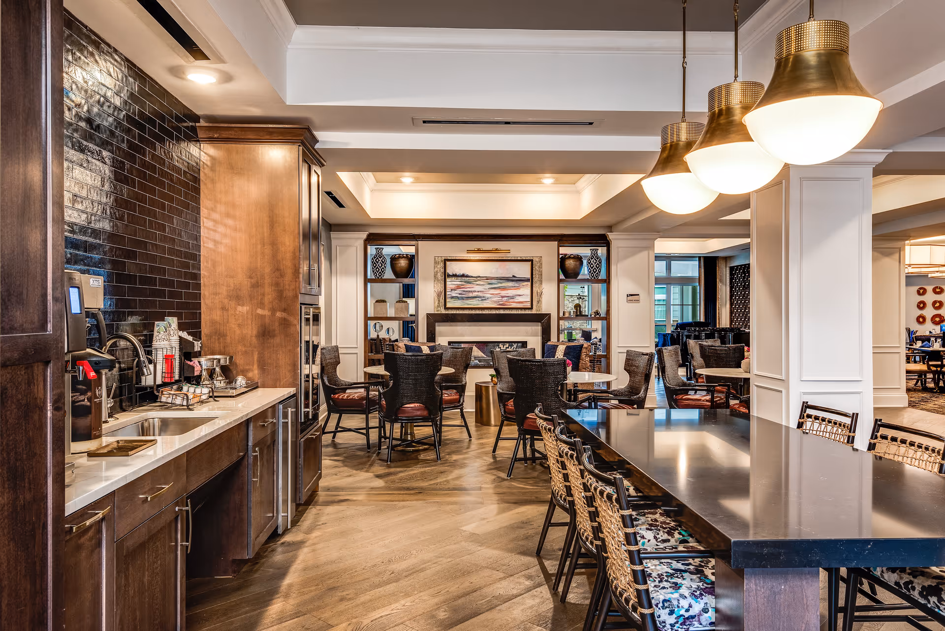 Interior view of a senior living facility common area featuring a kitchenette with dark wood cabinets and a sink on the left, a large black dining table with woven chairs on the right, and several round tables with cushioned chairs in the background. The room is well-lit with hanging brass pendant lights and recessed ceiling lights. Decorative vases and a framed painting are displayed on shelves around a fireplace in the background.