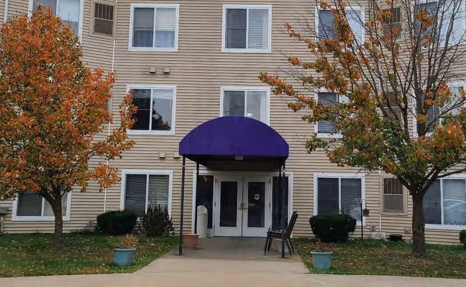Exterior view of a beige multi-story apartment building with a purple awning over the entrance. Two trees with autumn-colored leaves flank the entrance, and there are potted plants and stacked chairs near the doorway.