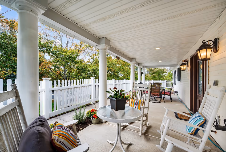 Covered outdoor porch area with white rocking chairs, a small round glass table with a potted plant, and a white picket fence surrounding the porch. The porch has ceiling lights and wall-mounted lantern-style lights, with trees visible in the background.