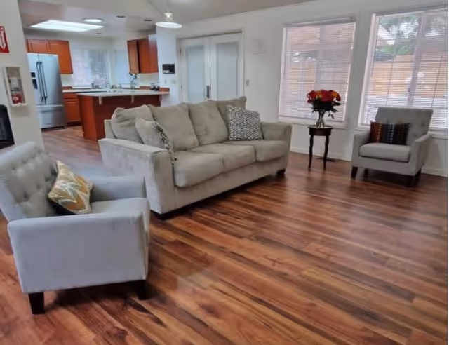 A spacious living room with wooden flooring featuring a beige sofa with patterned cushions, two gray armchairs with decorative pillows, a small wooden side table with a vase of red and orange flowers, and large windows with blinds allowing natural light. The background shows a kitchen area with wooden cabinets and stainless steel appliances.