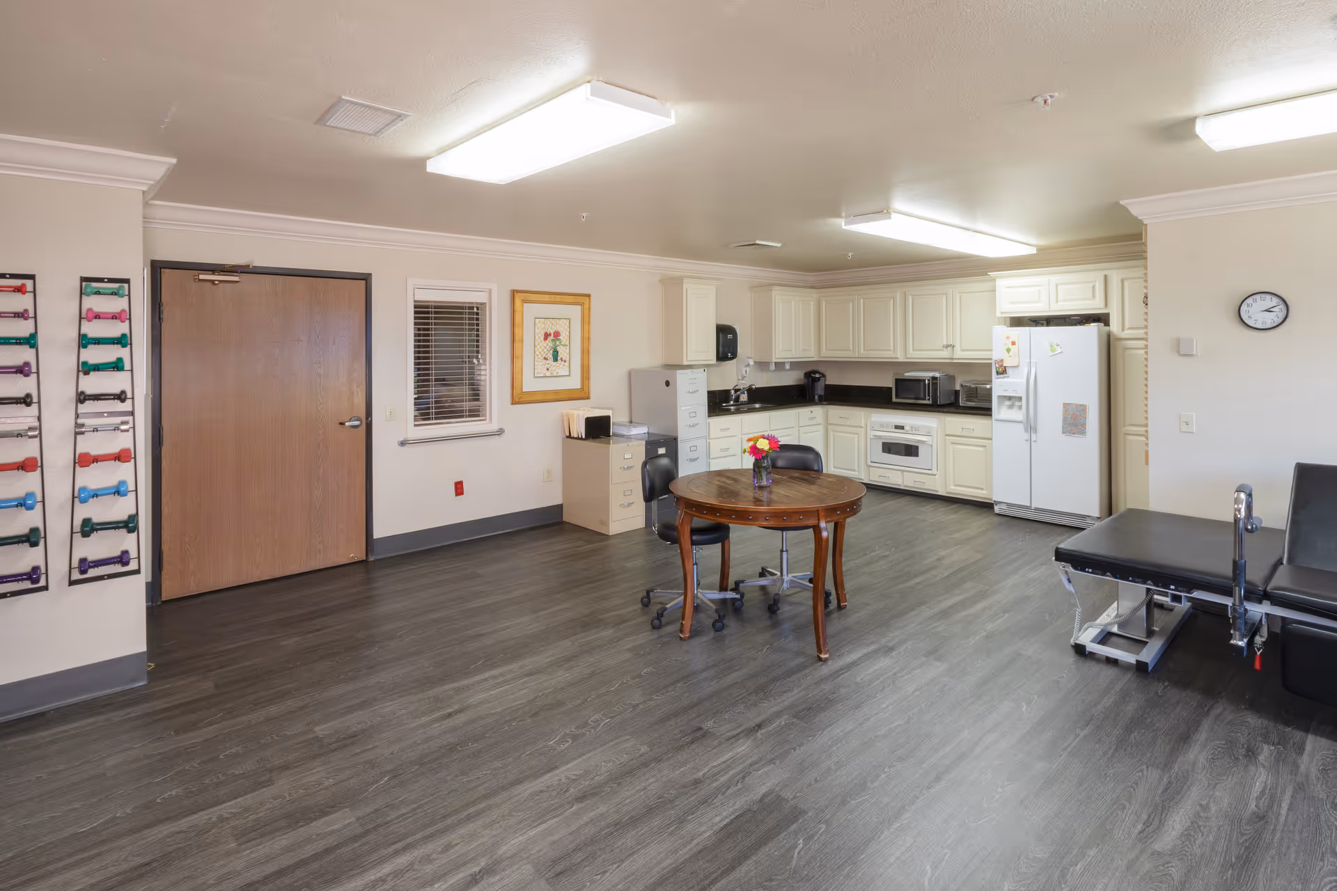 A spacious room with a kitchenette featuring white cabinets, a refrigerator, microwave, and stove. In the center, there is a round wooden table with a vase of flowers and two black office chairs on wheels. To the right, there is a black examination or treatment table. On the left wall, there is a rack holding colorful dumbbells, a wooden door, a small window with blinds, and a framed picture. The floor is dark wood, and the ceiling has bright fluorescent lights.