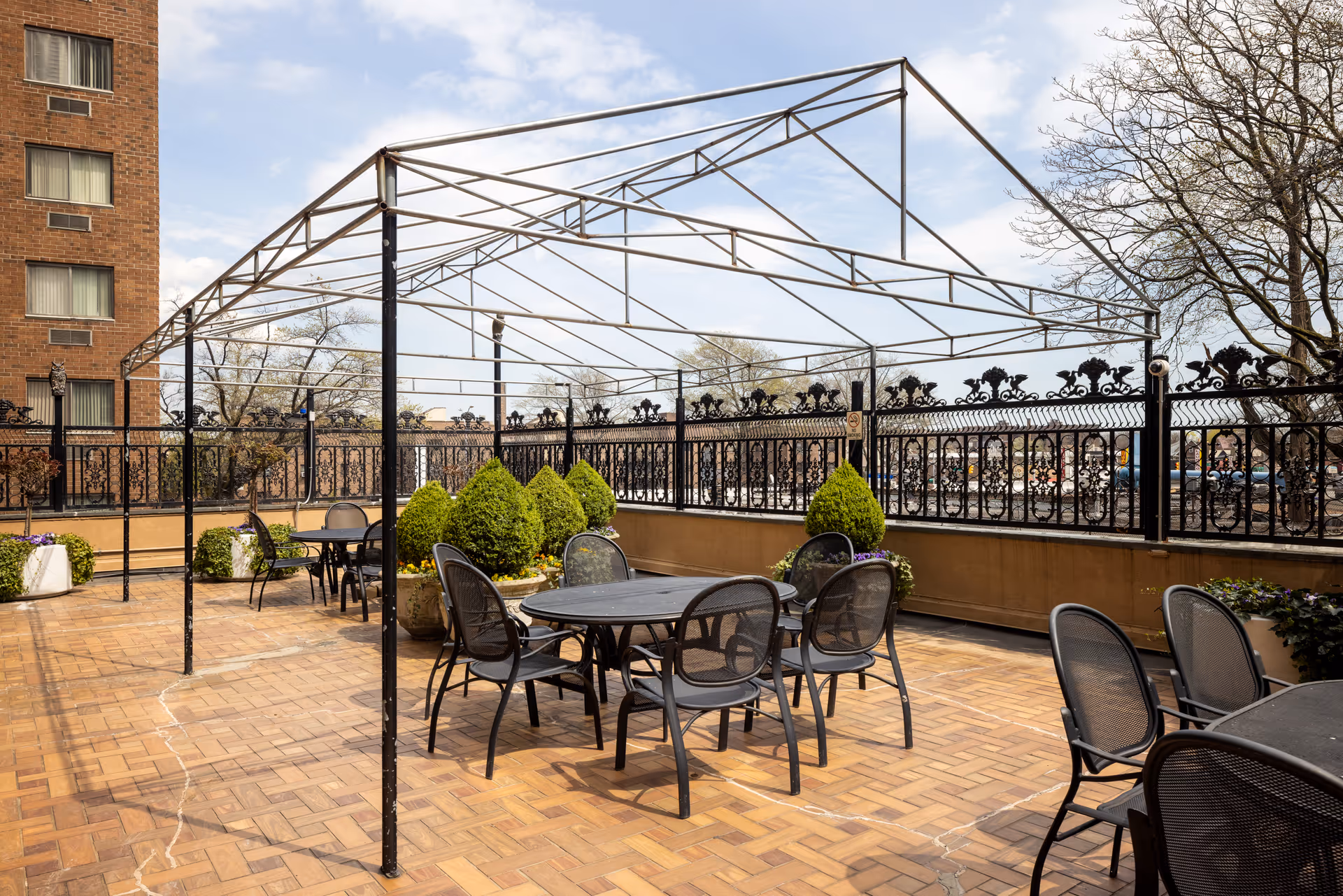 Outdoor rooftop patio with a metal pergola frame, round tables and chairs, potted shrubs, and a decorative wrought-iron railing.