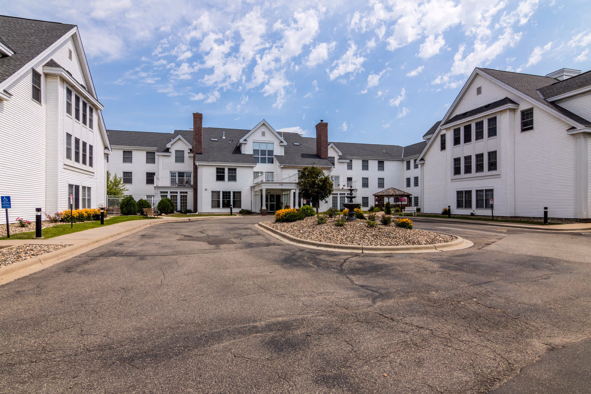 Exterior view of a senior living facility with white buildings surrounding a circular driveway with a landscaped island in the center. The sky is partly cloudy and the area is well-maintained with flowers and shrubs.
