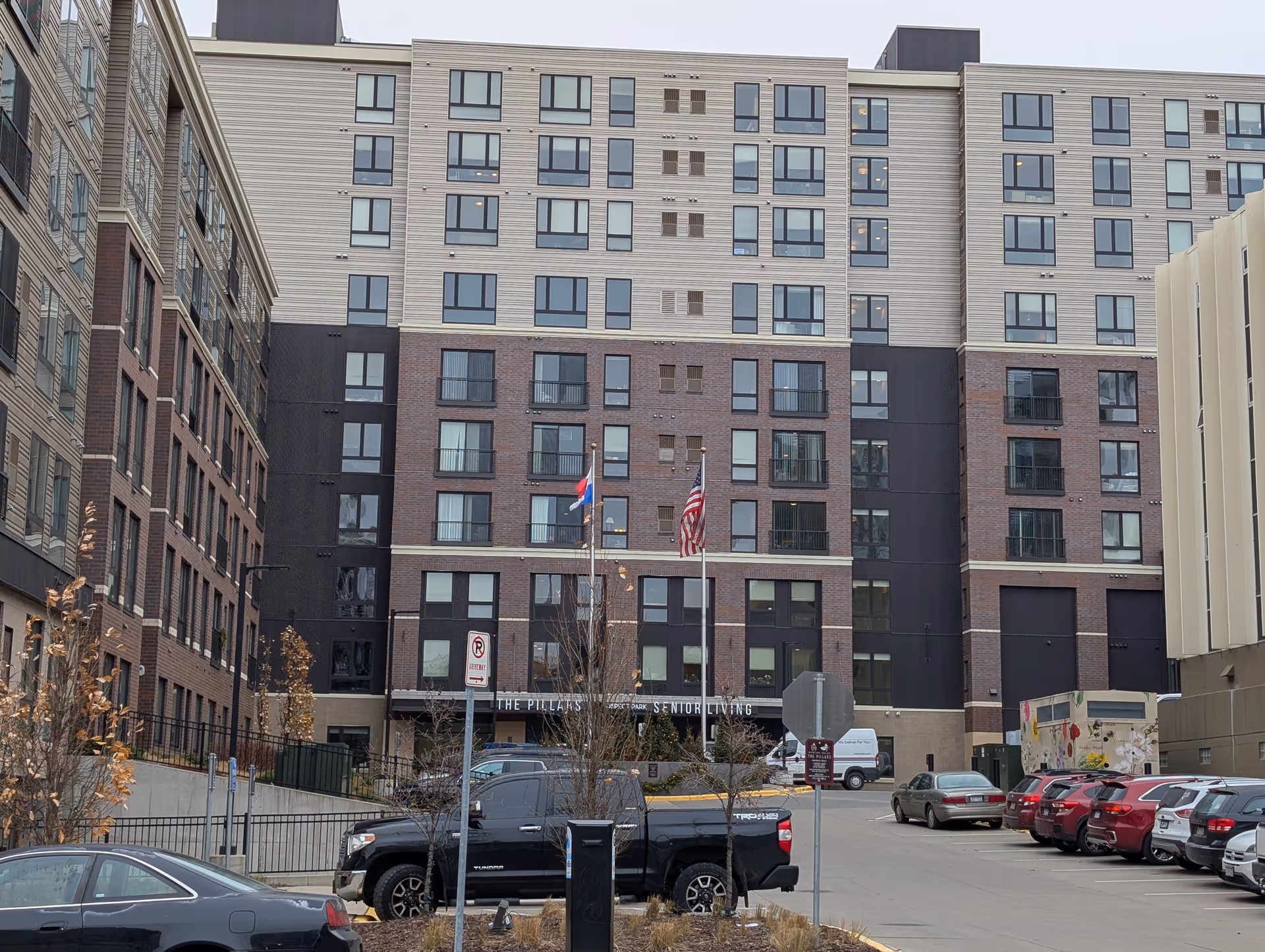 Front exterior of a multi-story senior living building with flags at the entrance and cars parked in the driveway.
