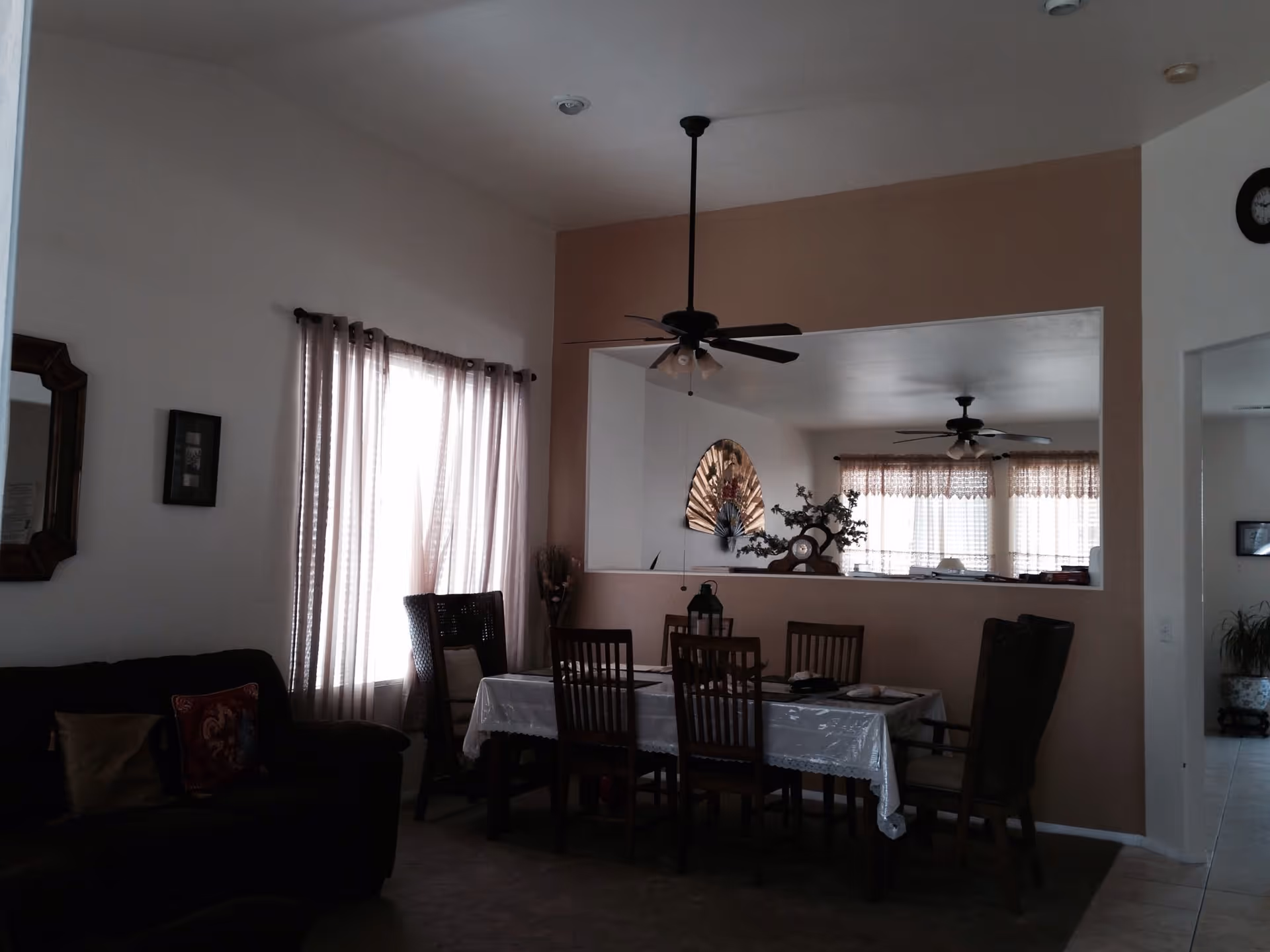 Interior view of a senior living facility showing a dining area with a table covered by a white tablecloth and six chairs around it. There is a ceiling fan above the table, a decorative fan on the wall, and a clock on the adjacent wall. Light filters through sheer curtains on the windows, and part of a living room with a dark sofa and cushions is visible on the left side.