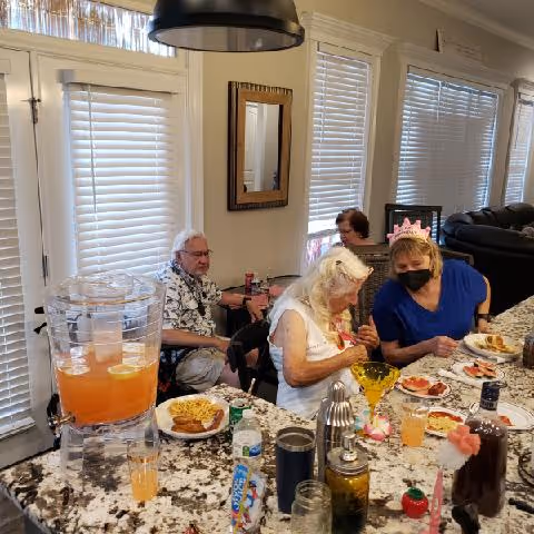 A group of elderly people sitting around a kitchen island with food and drinks. One woman is wearing a pink crown and a face mask. The kitchen has white blinds on the windows and a large black pendant light hanging above the island.