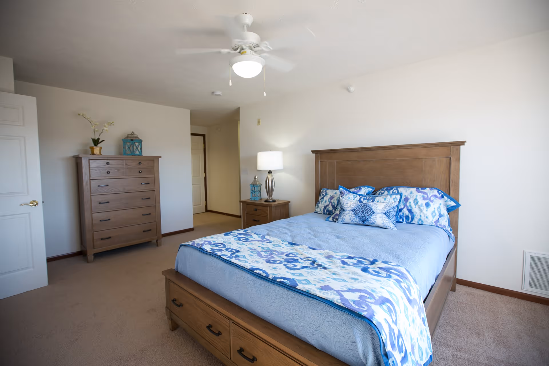 A bedroom in a retirement community featuring a wooden bed with blue and white patterned bedding and pillows. There is a wooden dresser with decorative items on top, a nightstand with a lamp and a decorative lantern, beige carpet, white walls, and a ceiling fan with a light.