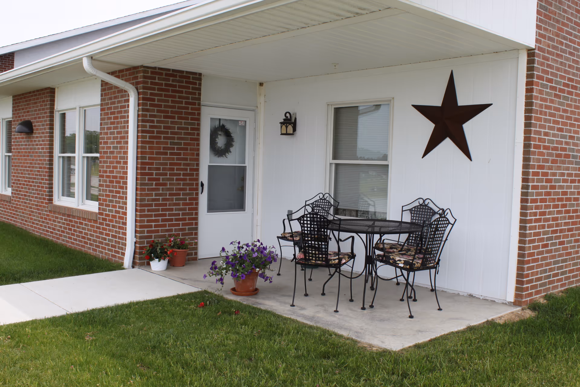 Outdoor patio area at Parkview Village Christian Care featuring a small round metal table with four matching chairs with floral cushions. The patio is concrete and adjacent to a brick and white siding building wall with a large decorative star mounted on it. There are potted plants with flowers near the door and on the patio. The door has a wreath hanging on it.