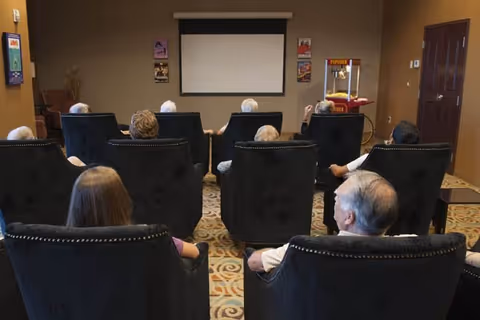 A group of elderly people seated in black armchairs facing a blank projector screen in a small theater room. There is a popcorn machine on the right side near the wall, and several movie posters are displayed on the wall behind the screen.