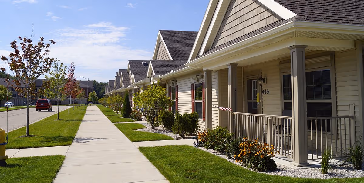 Row of single-story residential units with porches lining a sidewalk and manicured lawns.