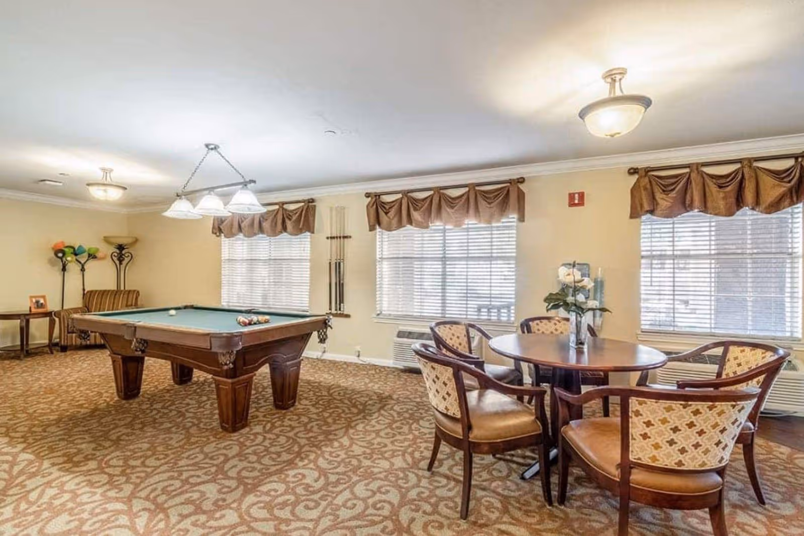 A well-lit recreational room featuring a pool table with balls arranged for a game, a round wooden table with four cushioned chairs, and large windows with brown valances letting in natural light. The room has patterned carpet flooring and beige walls, with a floor lamp and a small side table in the corner.