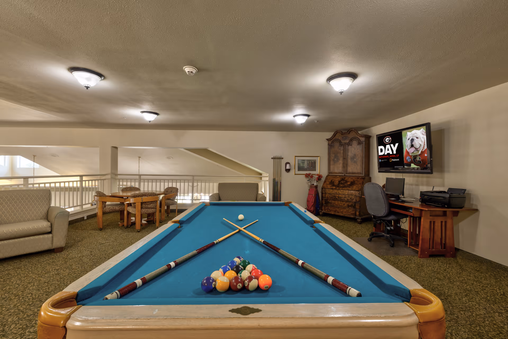 Interior view of a recreational room featuring a pool table with cues and balls arranged for a game. The room includes seating areas with sofas and a table with chairs. A desk with a computer and printer is positioned against the wall, and a TV mounted on the wall displays an image of a bulldog with the text 'DAY'. The room has carpeted floors and ceiling lights.