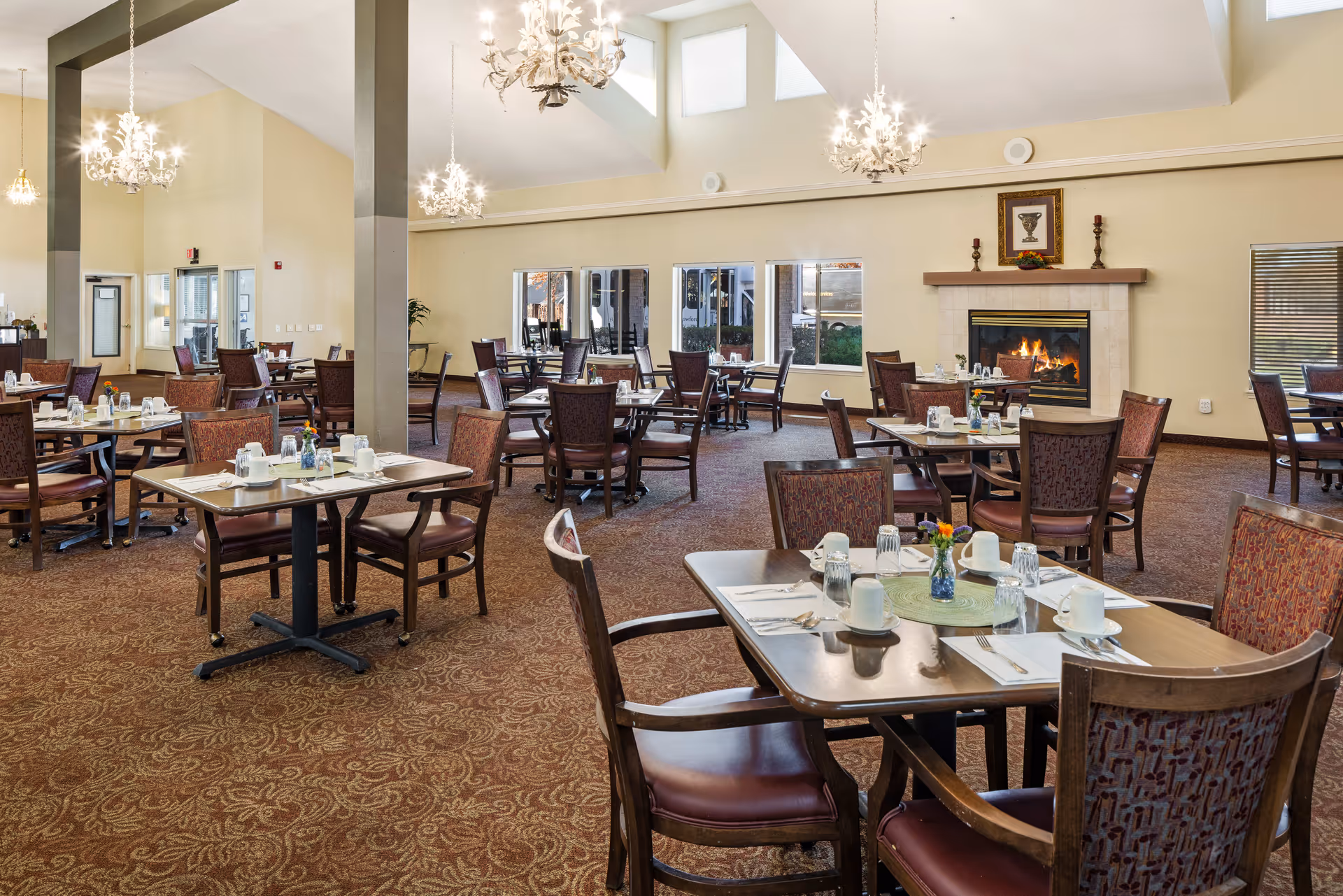A spacious dining room in a senior living community with multiple tables and chairs arranged neatly. Each table is set with white cups, glasses, napkins, and silverware. The room features a patterned carpet, large windows letting in natural light, and a lit fireplace with decorative items on the mantel. Elegant chandeliers hang from the ceiling, adding a warm ambiance.