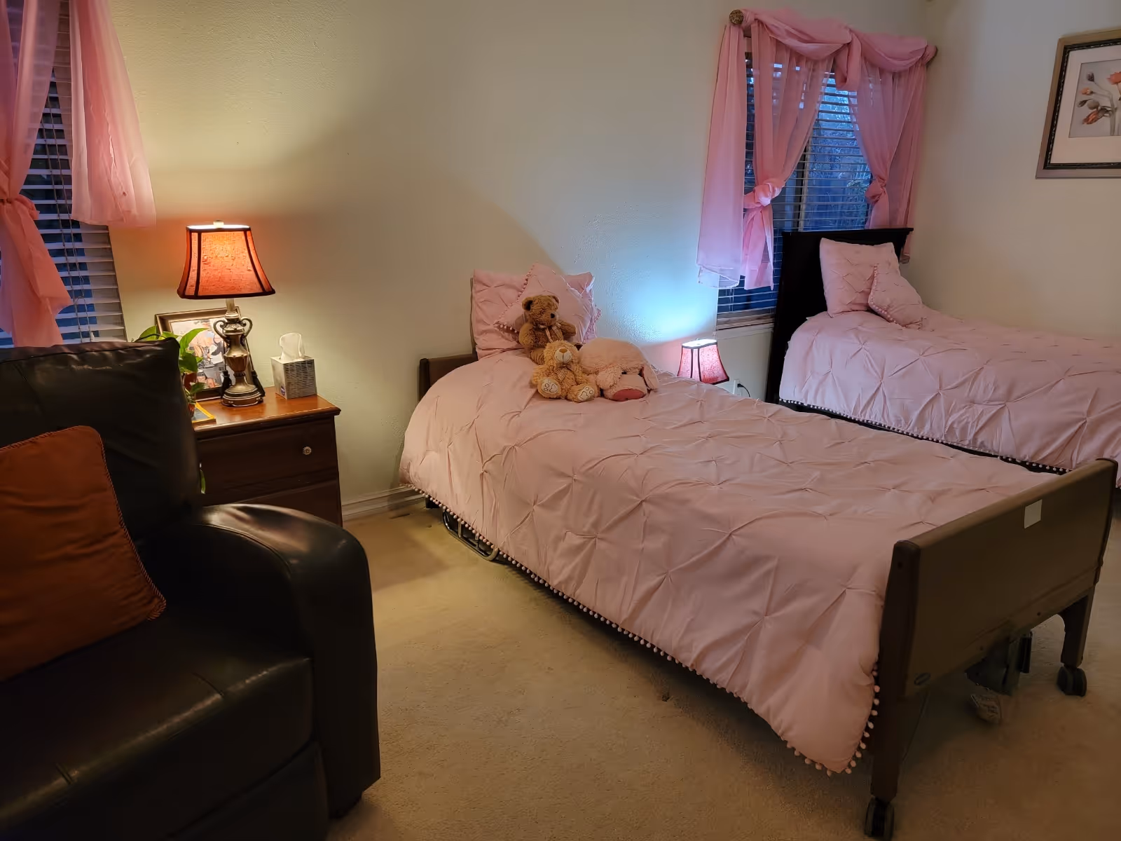 A softly lit bedroom with two twin beds dressed in pink bedding, a nightstand with a lamp, an armchair, and stuffed animals on one bed.